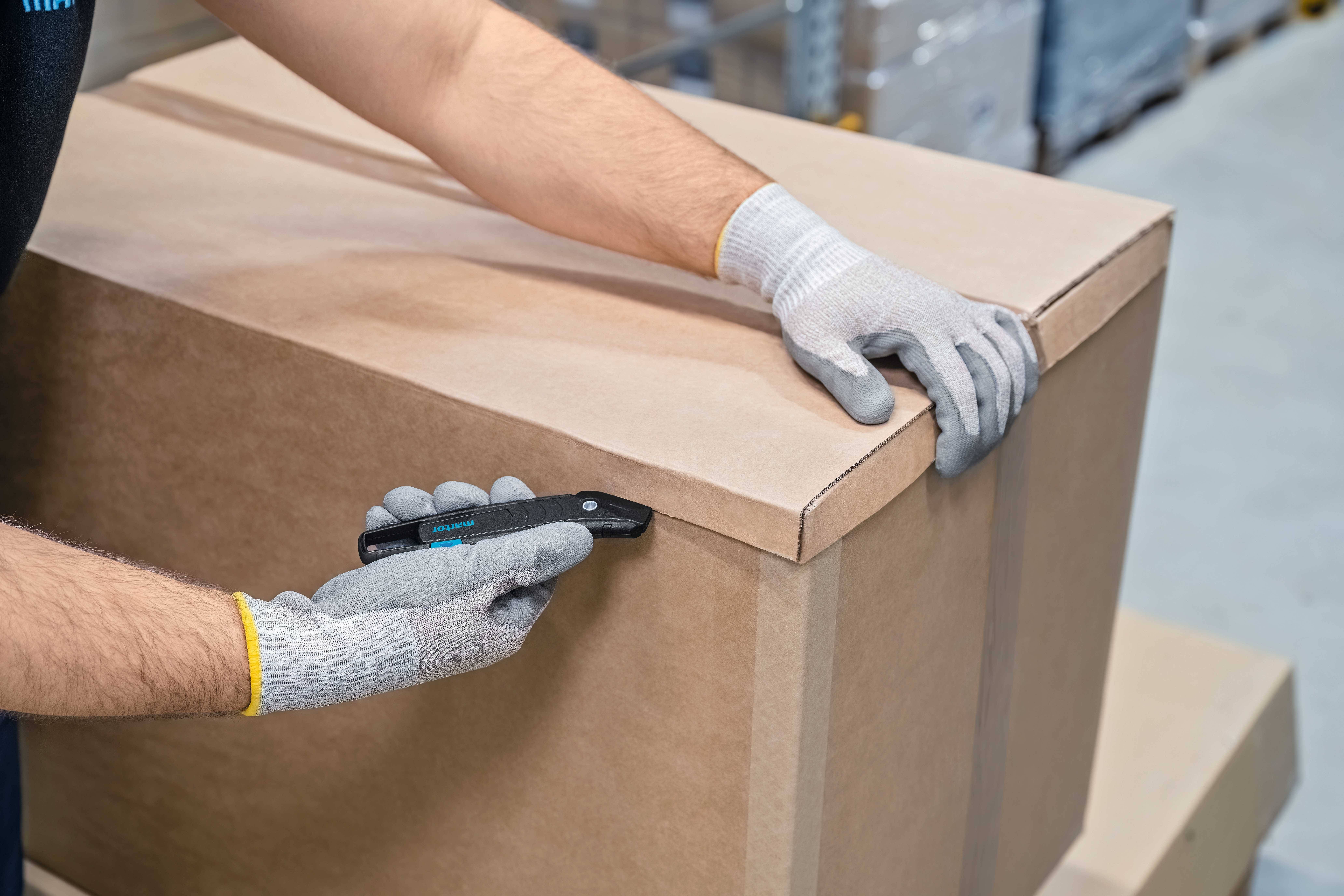 Eine Person mit grauen Handschuhen öffnet vorsichtig einen großen Karton mit einem schwarzen Sicherheitsmesser in einer Lagerumgebung.