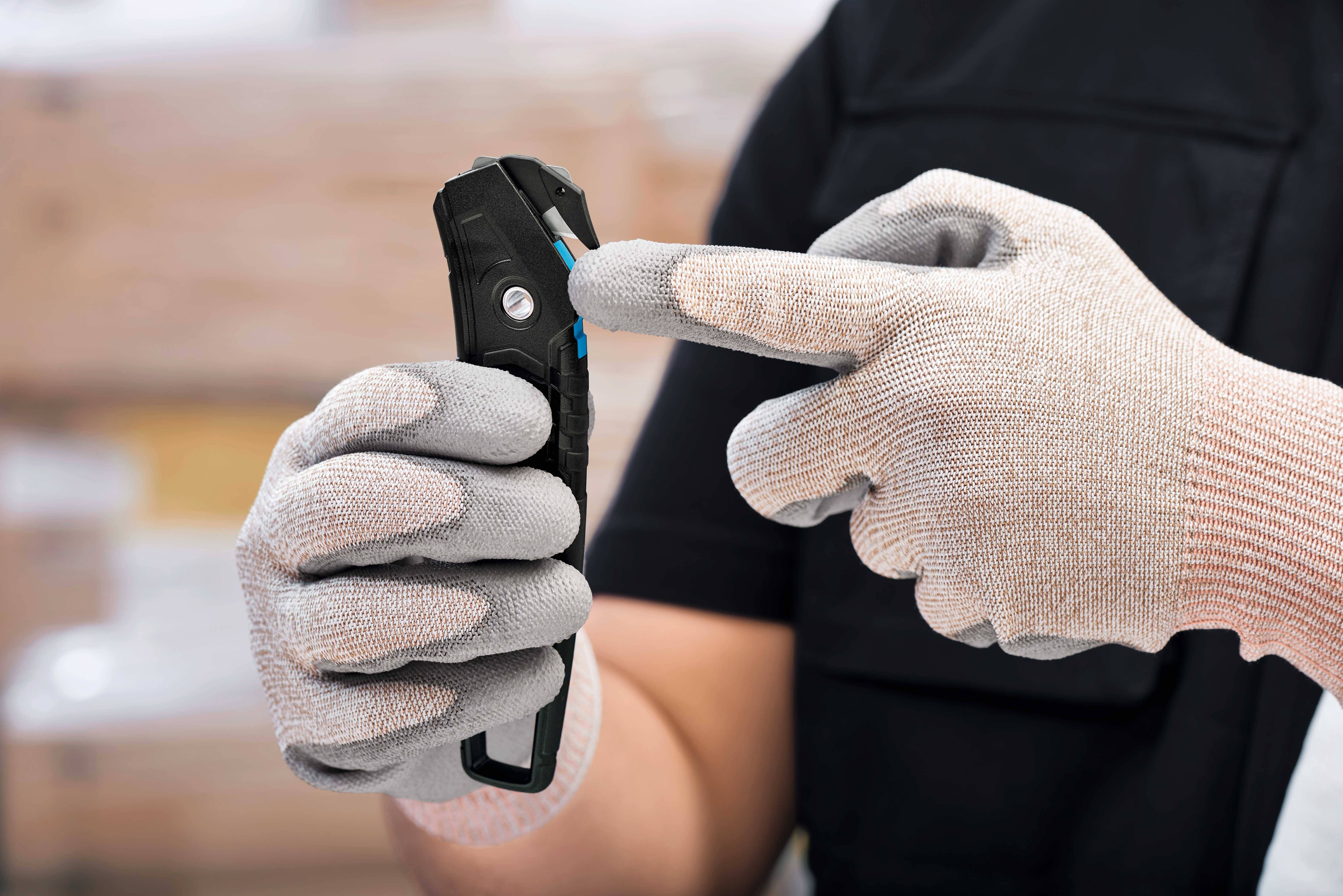 Eine Person mit grauen Sicherheitshandschuhen hält und benutzt ein schwarzes Sicherheitsmesser. Die Szene findet in einem industriellen Kontext statt.