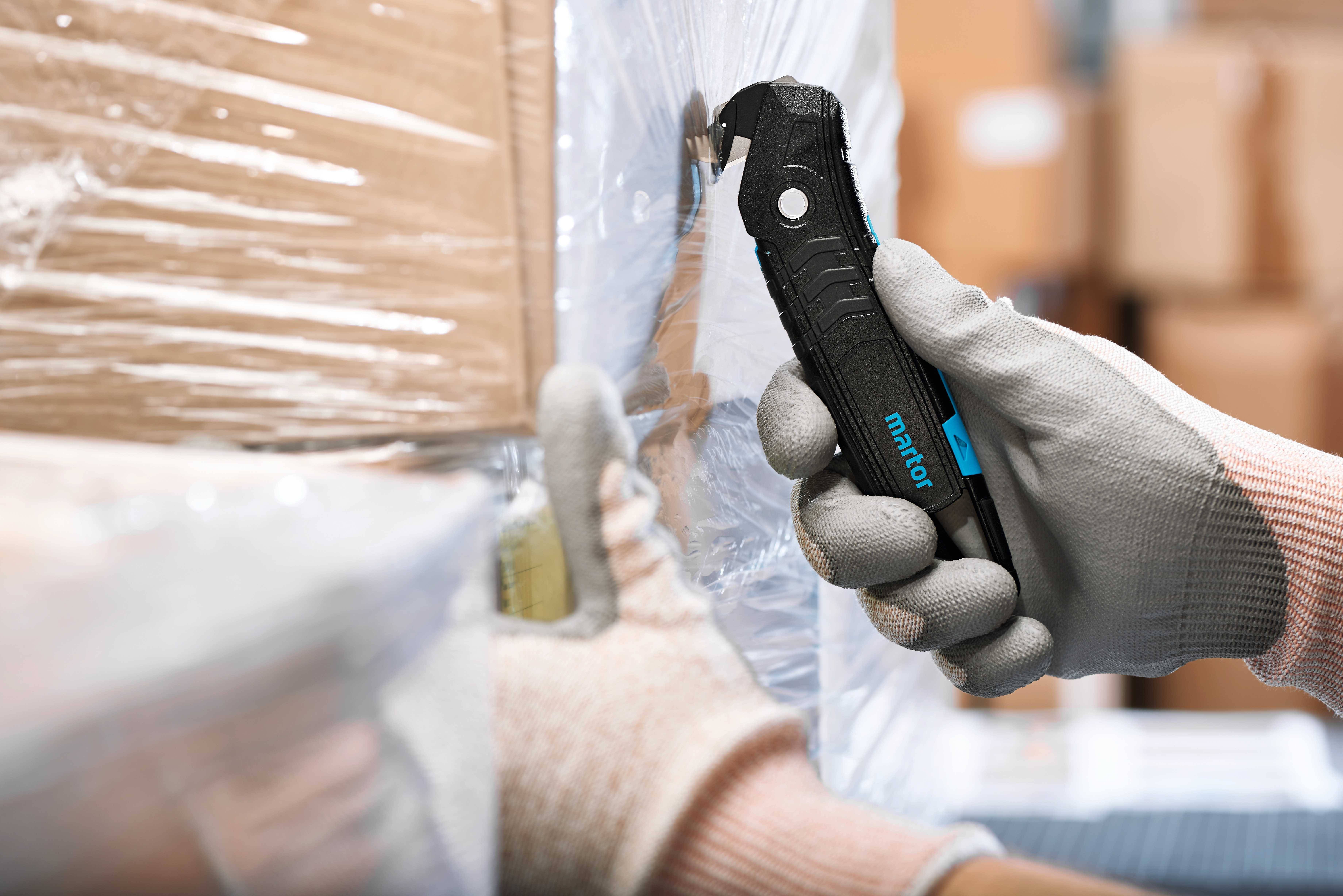 Eine Hand mit grauem Handschuh hält ein schwarzes Sicherheitsmesser und schneidet durch Verpackungsfolie um Kartons.