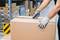 Eine Person öffnet mit einem Cutter ein großes Paket in einem Lagerhaus. Regale mit Kartons im Hintergrund sind erkennbar.