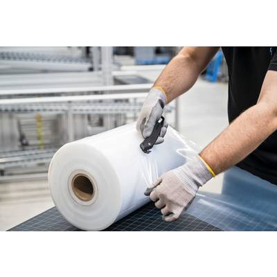 Eine Person schneidet Plastikfolie von einer großen Rolle mit einem Messer ab. Im Hintergrund sind industrielle Regale sichtbar.