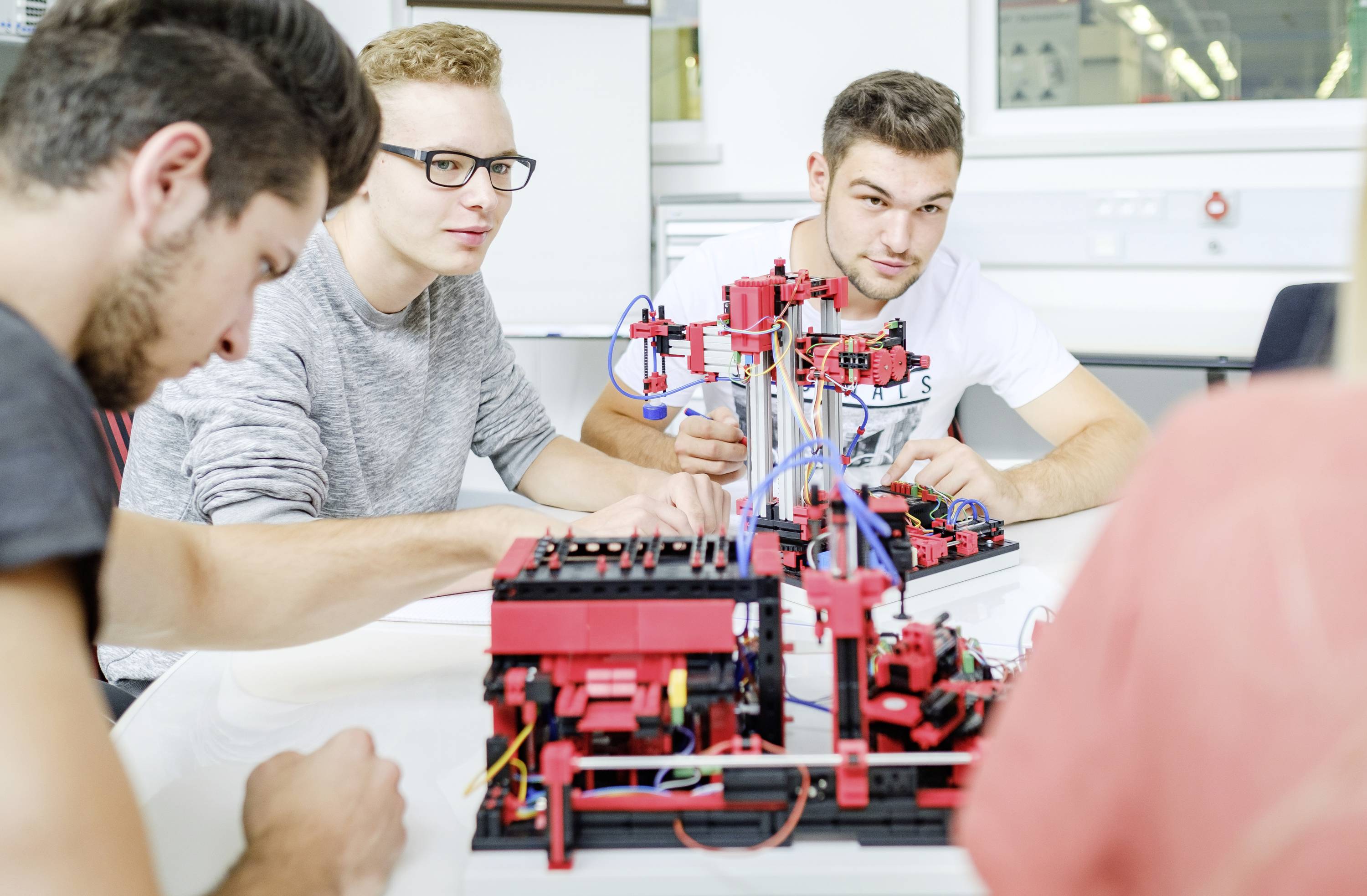 Vier junge Erwachsene arbeiten konzentriert an einem Tisch mit komplexen mechanischen Modellen und Elektronik, in einem Laborraum.