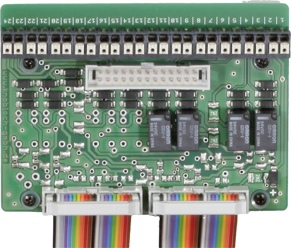 Eine grüne Leiterplatte mit zwei Reihen schwarzer Anschlüsse oben, mehreren schwarzen und silbernen Bauteilen, und zwei mehrfarbigen Kabeln unten.