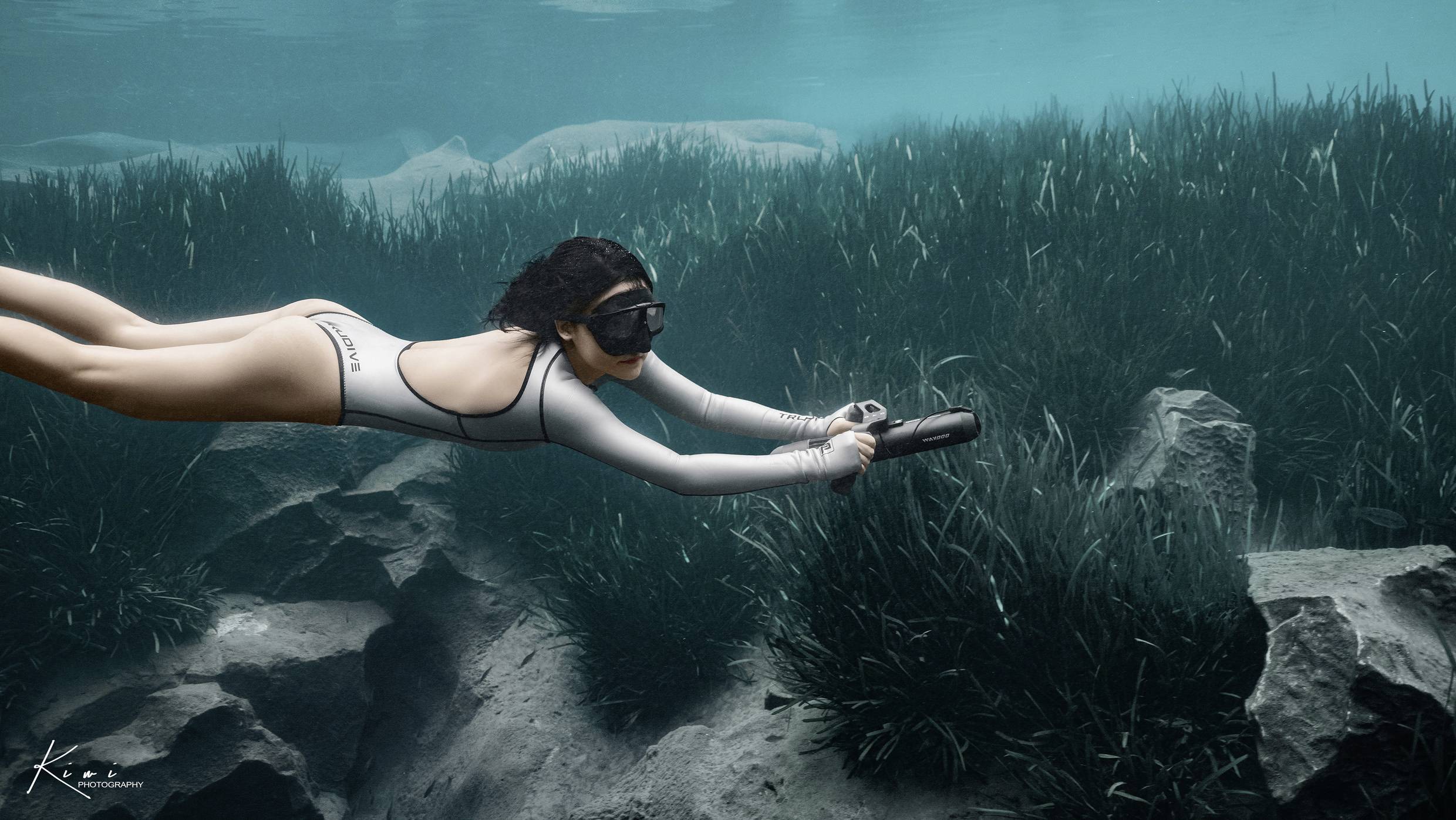 Eine Person taucht tief unter Wasser, in einem klaren, blauen Meer, trägt eine Taucherbrille und einen einteiligen Anzug, schwimmt über Seegras.