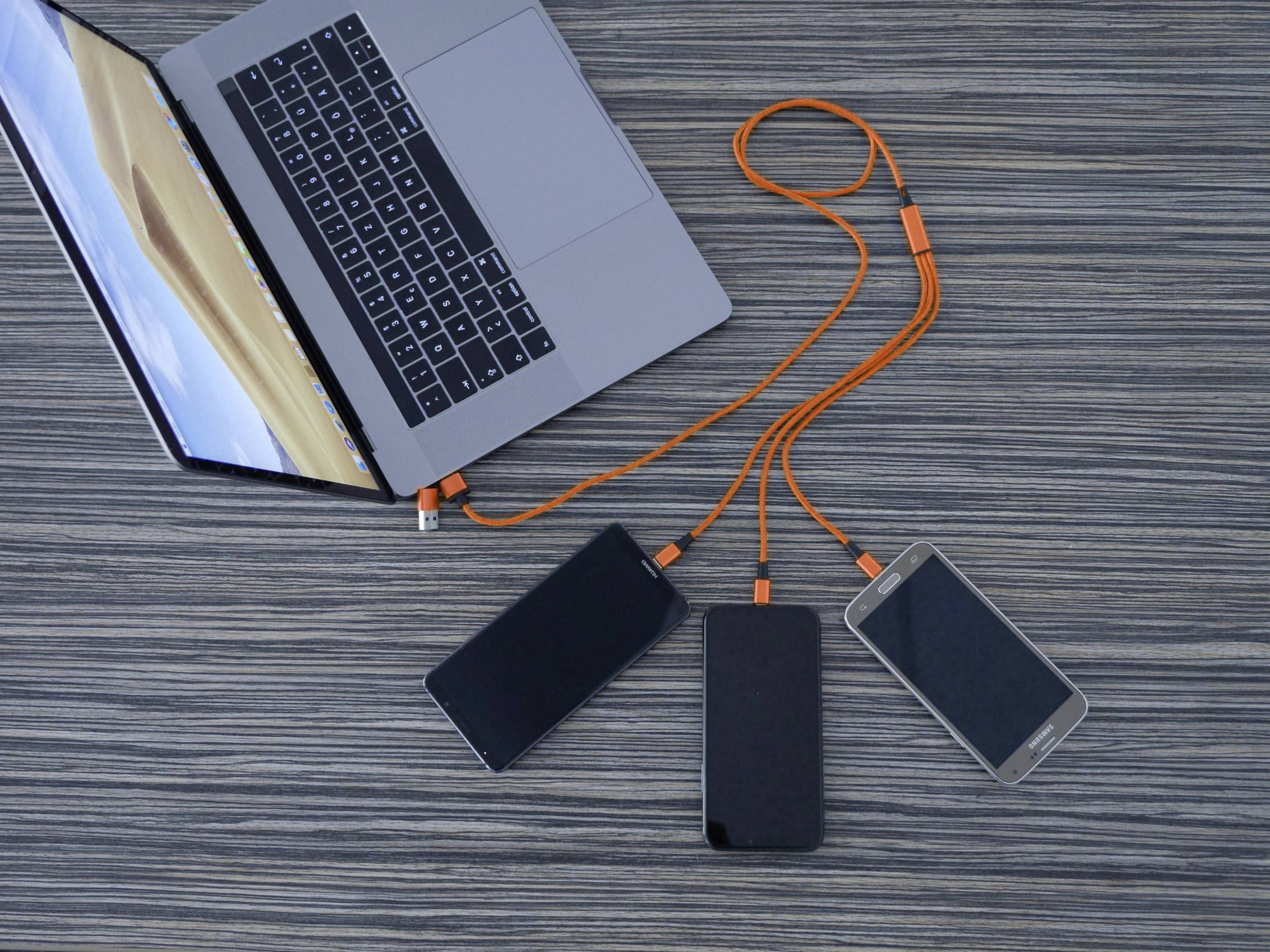 Ein Laptop ist mit drei Smartphones über orangefarbene Kabel verbunden, die auf einem Holztisch liegen.