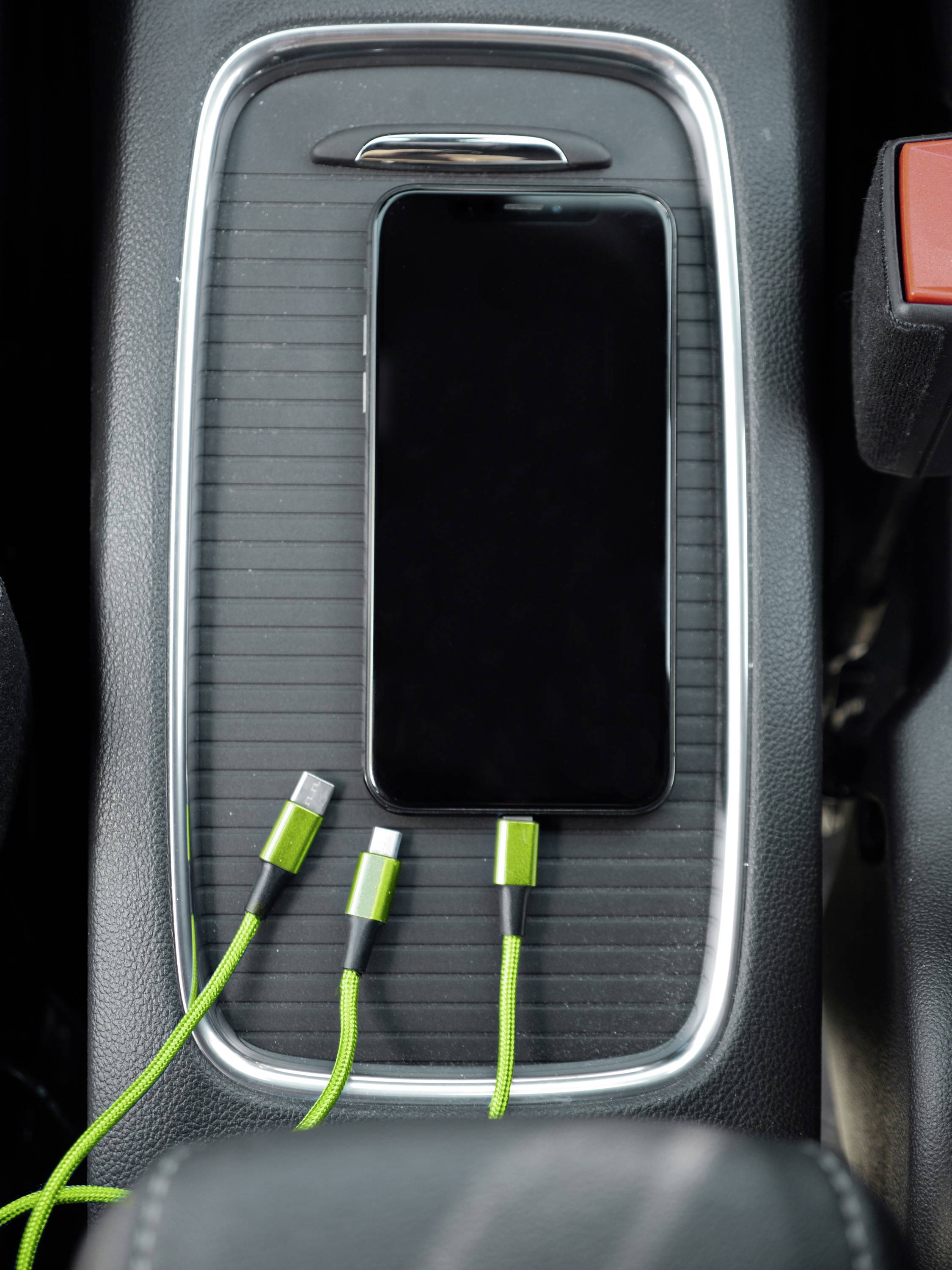 Ein schwarzes Smartphone liegt auf einer Armlehne im Auto, daneben ein grünes Ladekabel mit mehreren Anschlüssen.