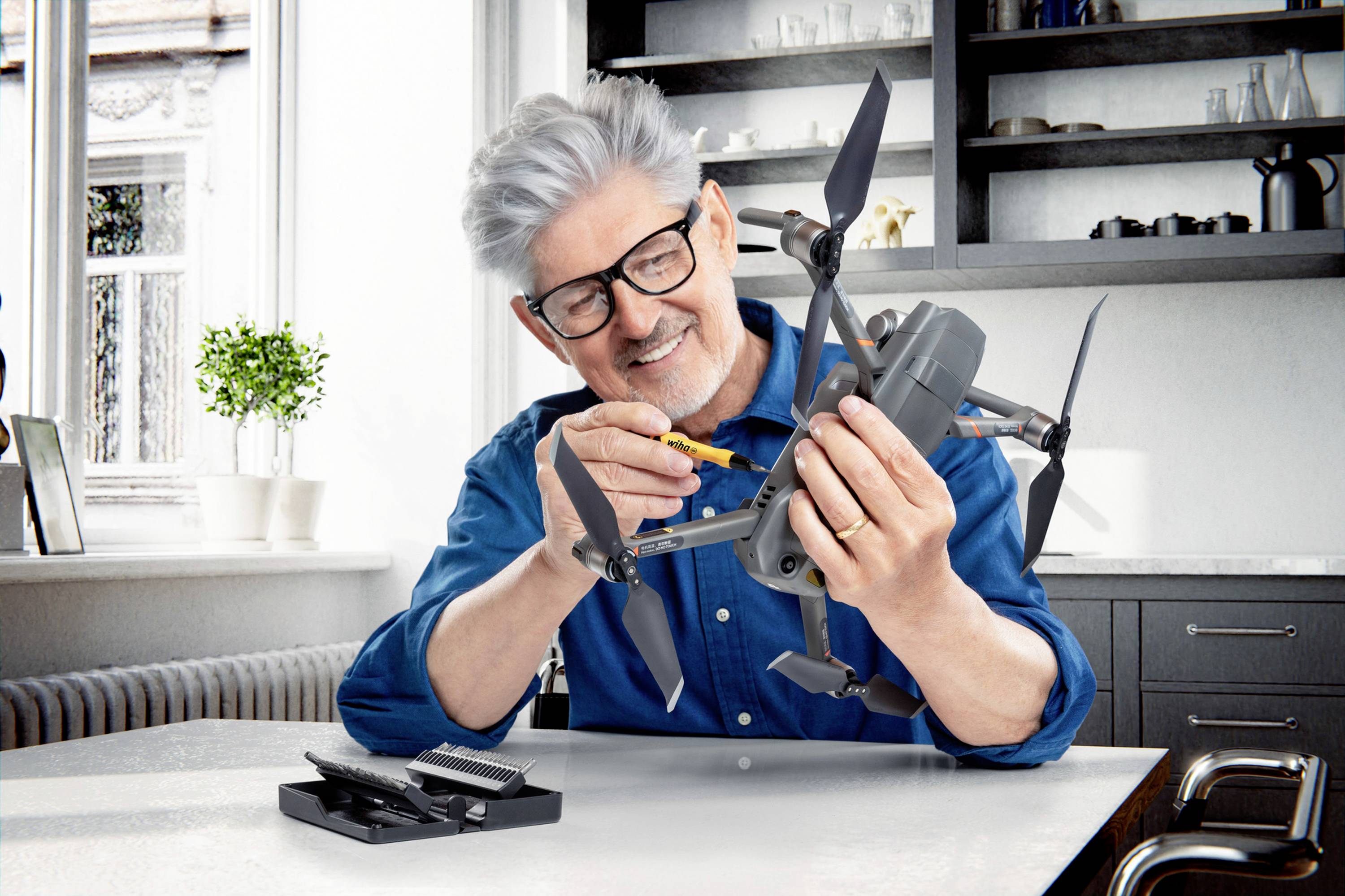 Ein älterer Mann mit Brille repariert lächelnd eine Drohne in einem modernen Raum. Auf dem Tisch liegen Werkzeuge.