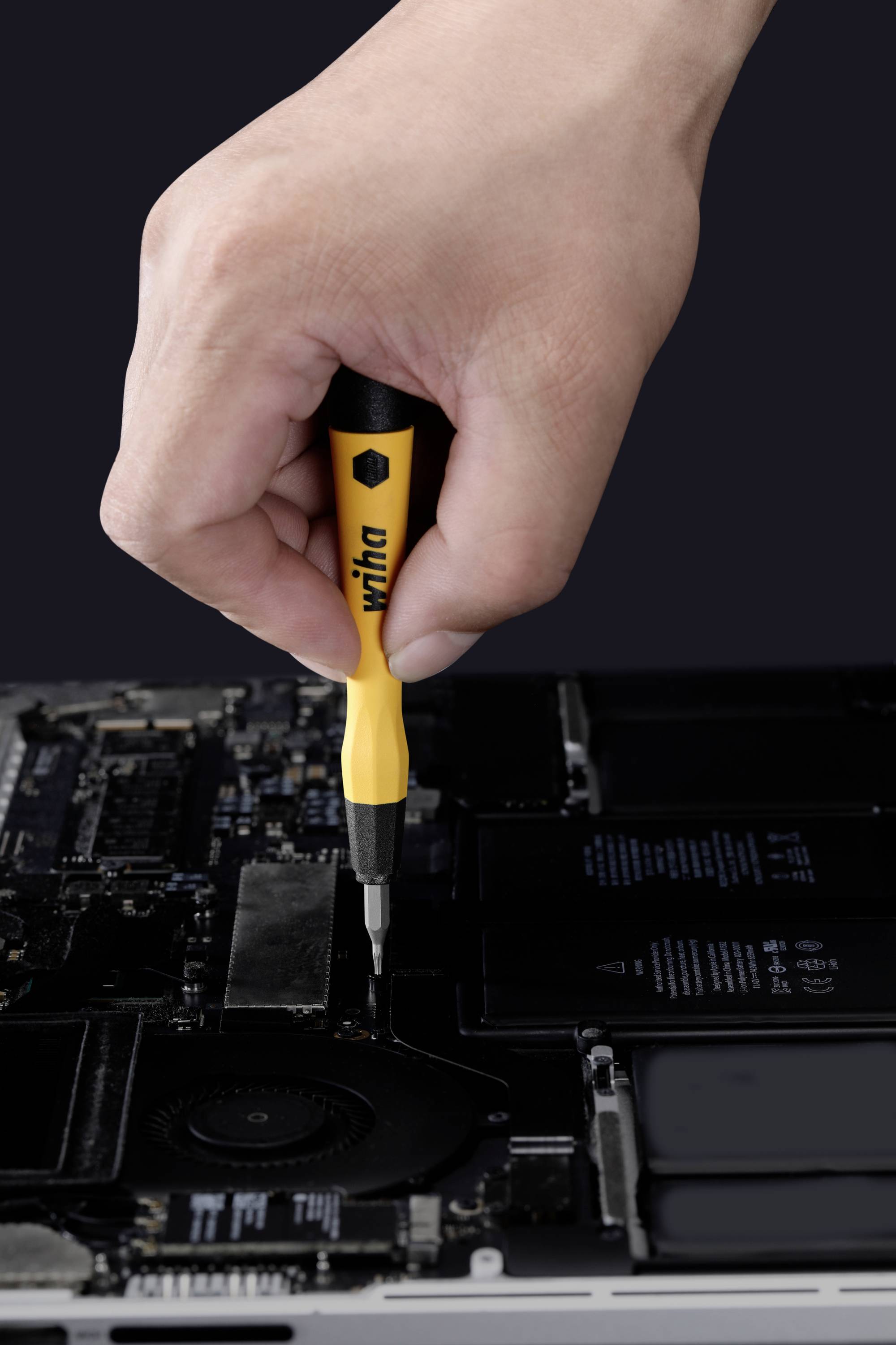 Eine Hand hält einen gelben Schraubendreher und schraubt an einem offenen Laptop. Komponenten und Batterie sind sichtbar.