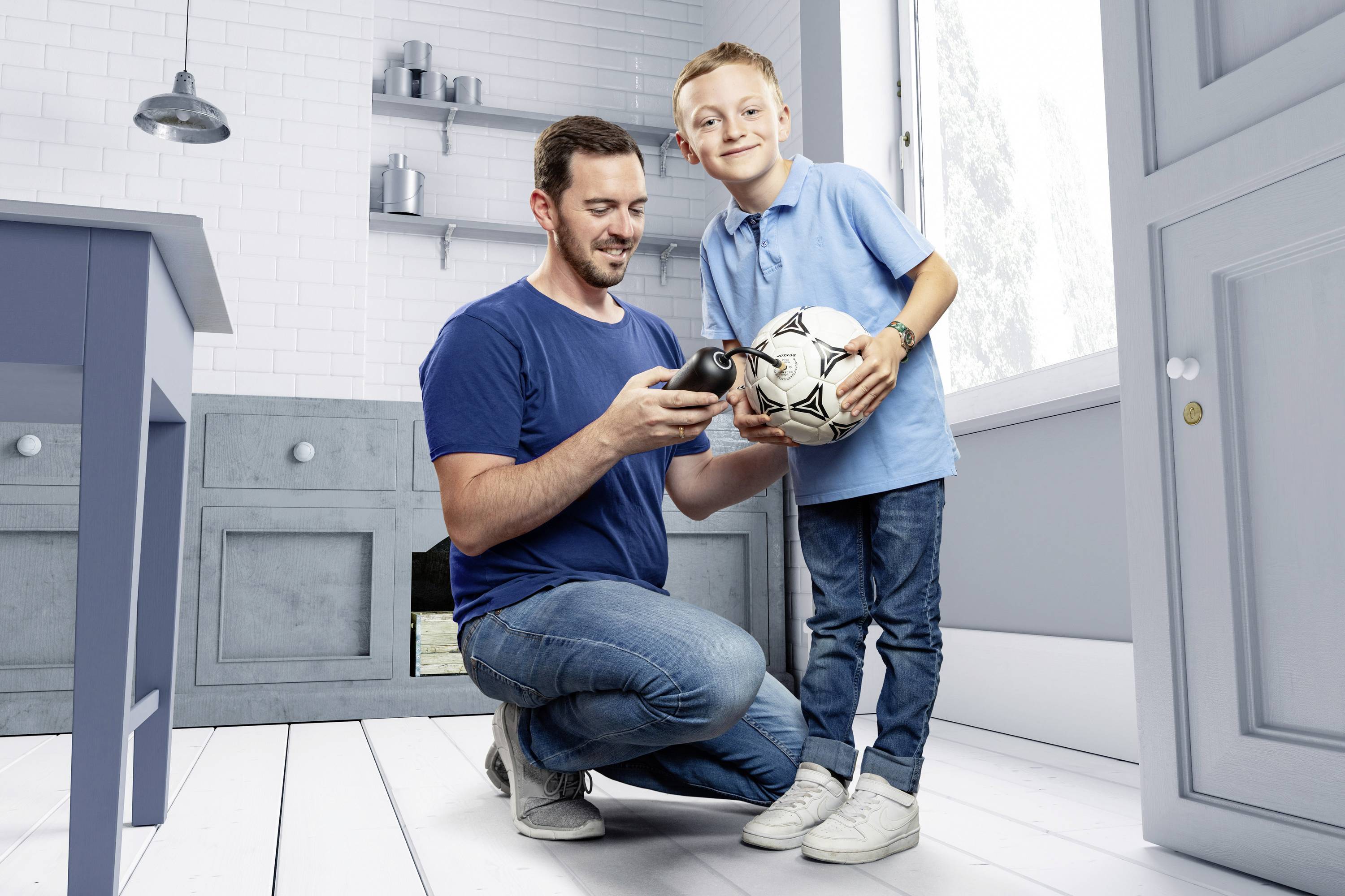 Ein Mann kniet neben einem Kind, das einen Fußball hält. Sie lächeln sich an in einem modernen, hellen Raum.