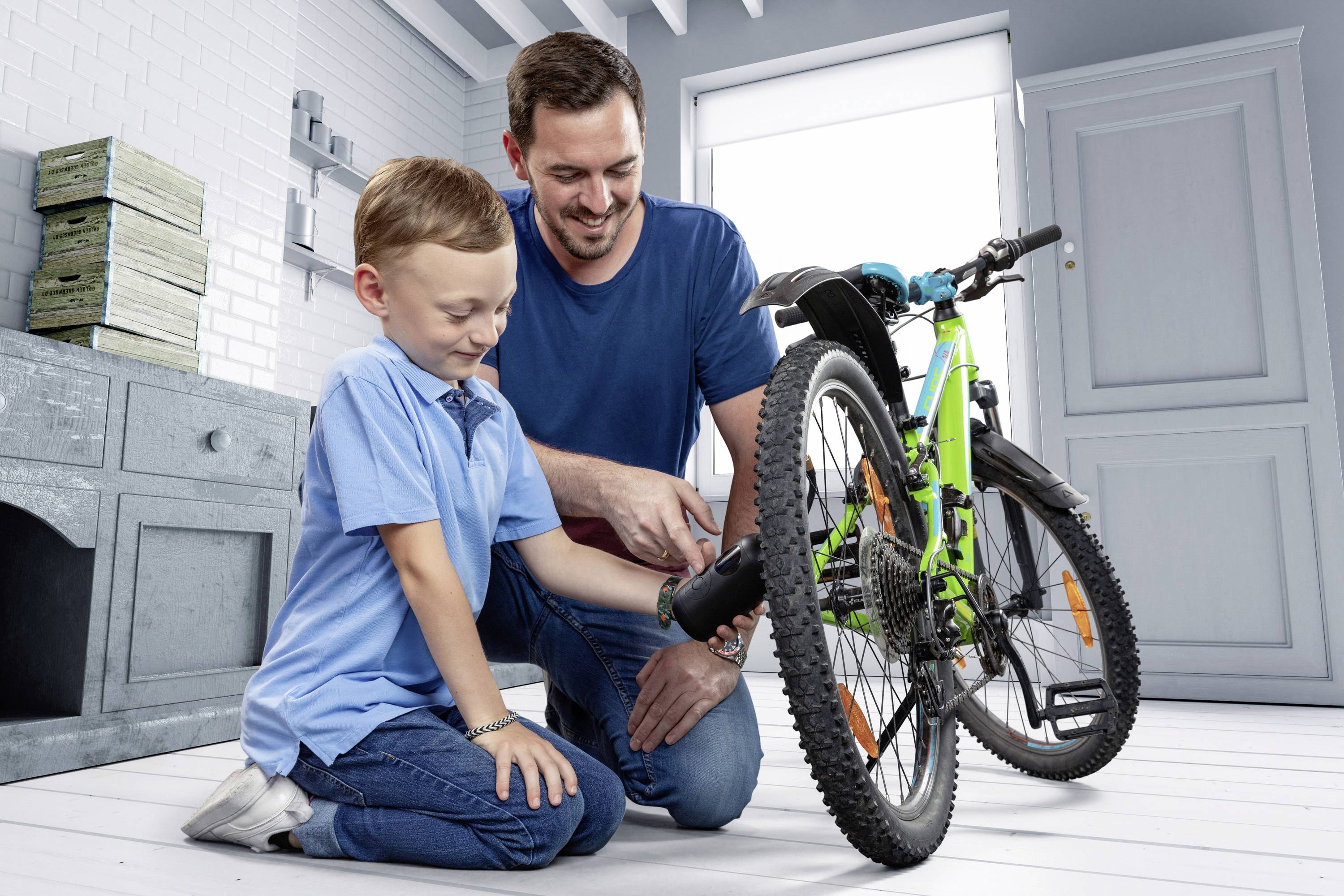 Ein Mann und ein Junge sitzen in einem hellen Raum und reparieren gemeinsam ein grünes Fahrrad. Der Mann zeigt dem Jungen etwas.