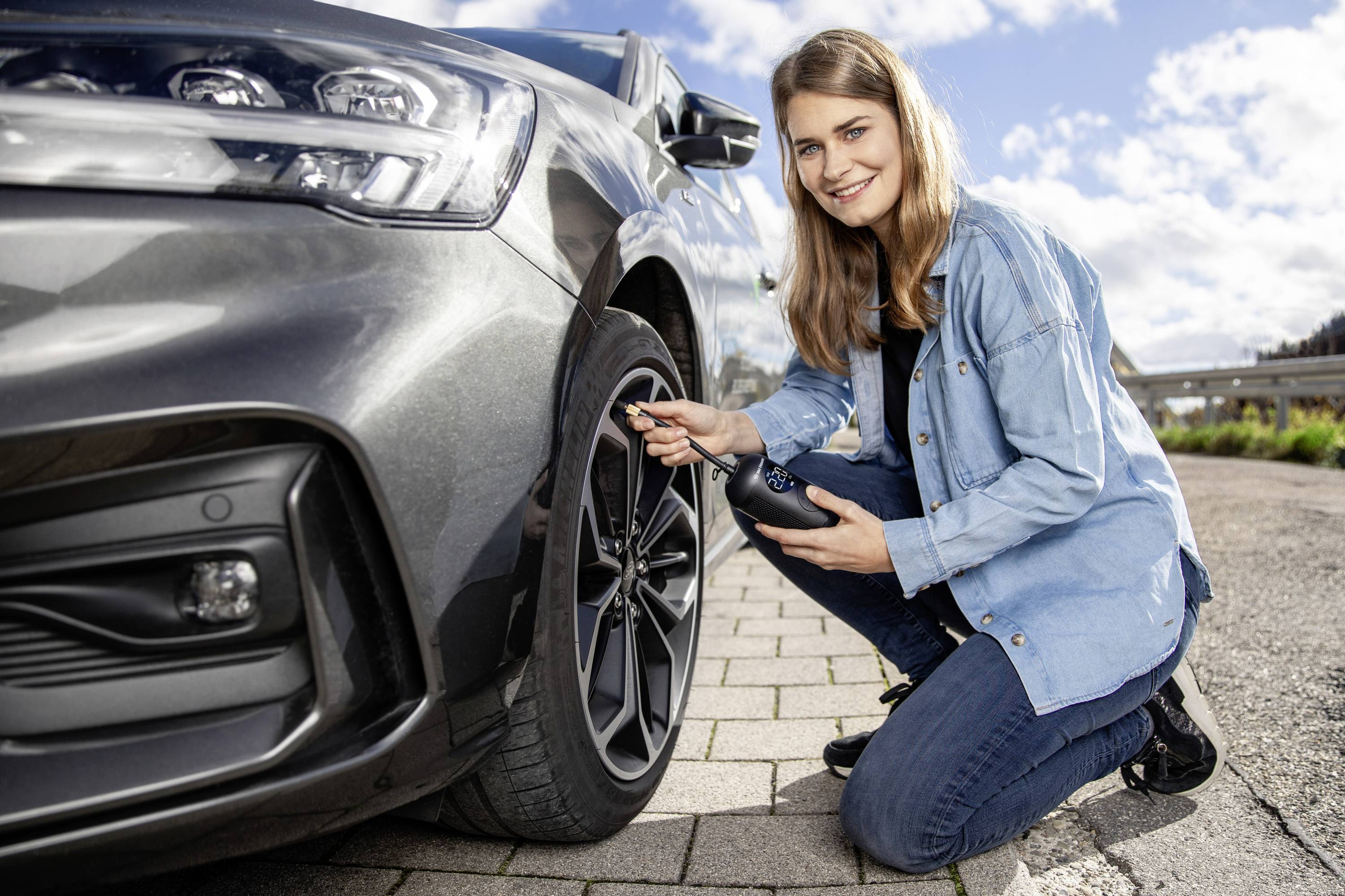 Eine Frau kniet neben einem Auto und überprüft den Reifendruck mit einem Messgerät. Der Himmel ist blau mit wenigen Wolken.