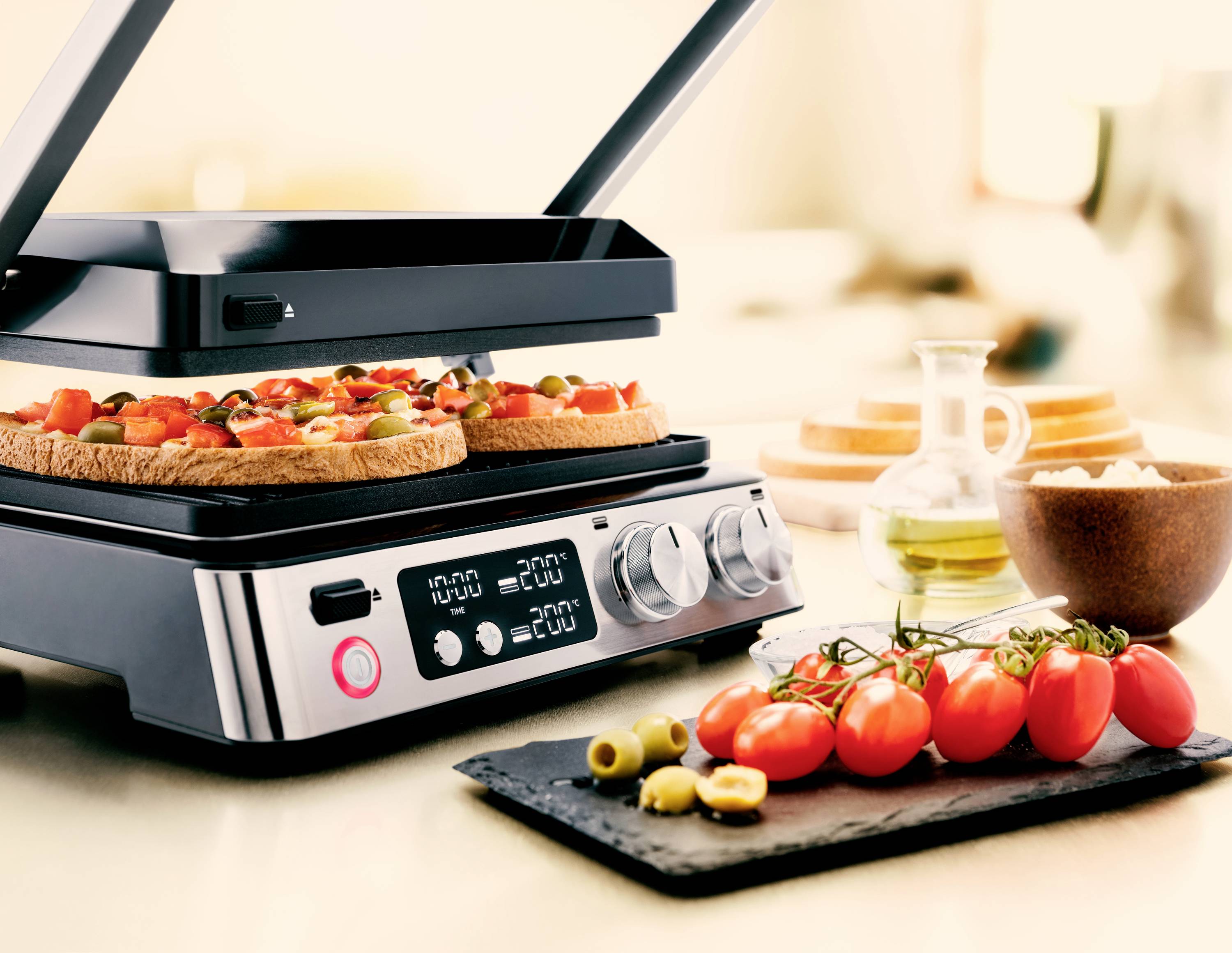 Ein moderner Elektrogrill auf einer Küchentheke grillt belegte Brotscheiben. Daneben liegen Tomaten, Oliven und eine Ölflasche.