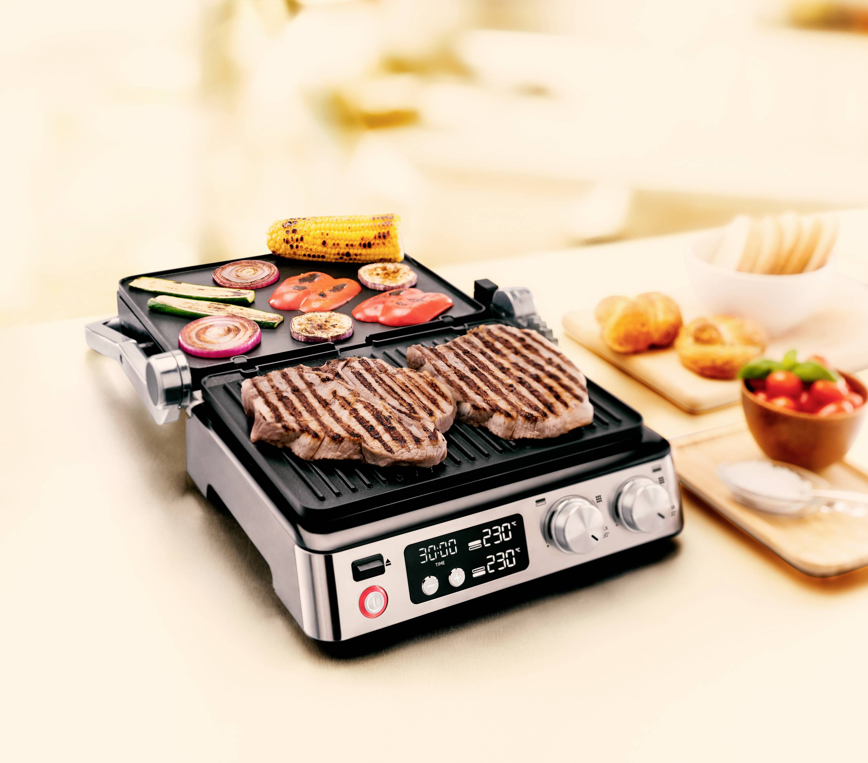 Ein elektrischer Grill mit zwei Fleischstücken auf der Grillplatte, daneben Gemüse wie Mais, Tomaten und Zucchini. In der Nähe stehen Brotkörbchen und eine Schüssel Salat.