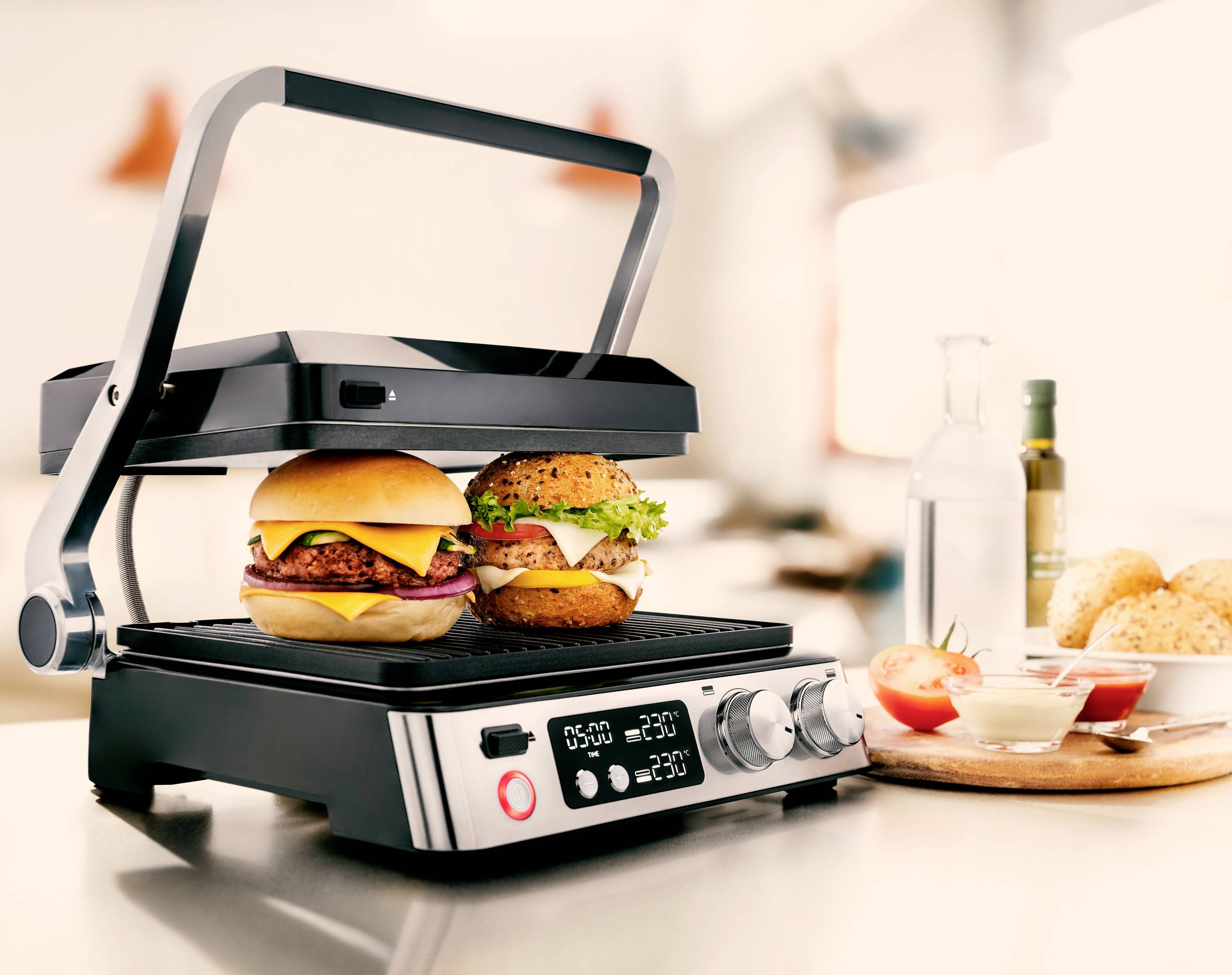 Elektrischer Grill in moderner Küche bereitet zwei Burger zu. Einer hat Rindfleisch, Käsescheiben und Salat, der andere Hähnchen und Salat.