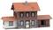 Ein Modellhaus mit orangem Dach, heller Fassade und braunen Fensterläden. Links Anbau, rechts Anbau mit offenem Eingang.