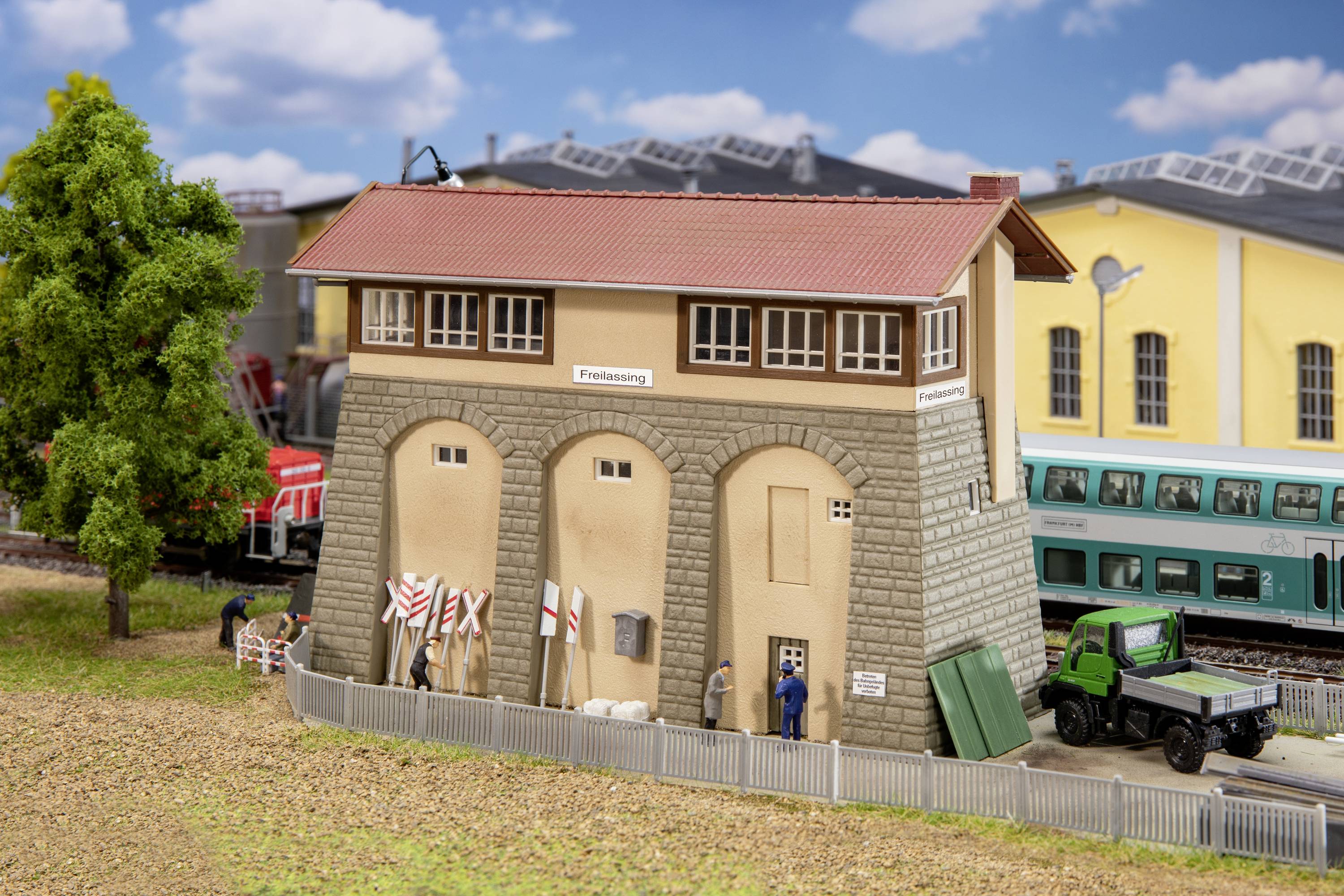Gebäude in Miniaturlandschaft: Das Stellwerk 'Freilassing' mit roten Ziegeldach, daneben Züge und ein grüner LKW, umgeben von Bäumen.