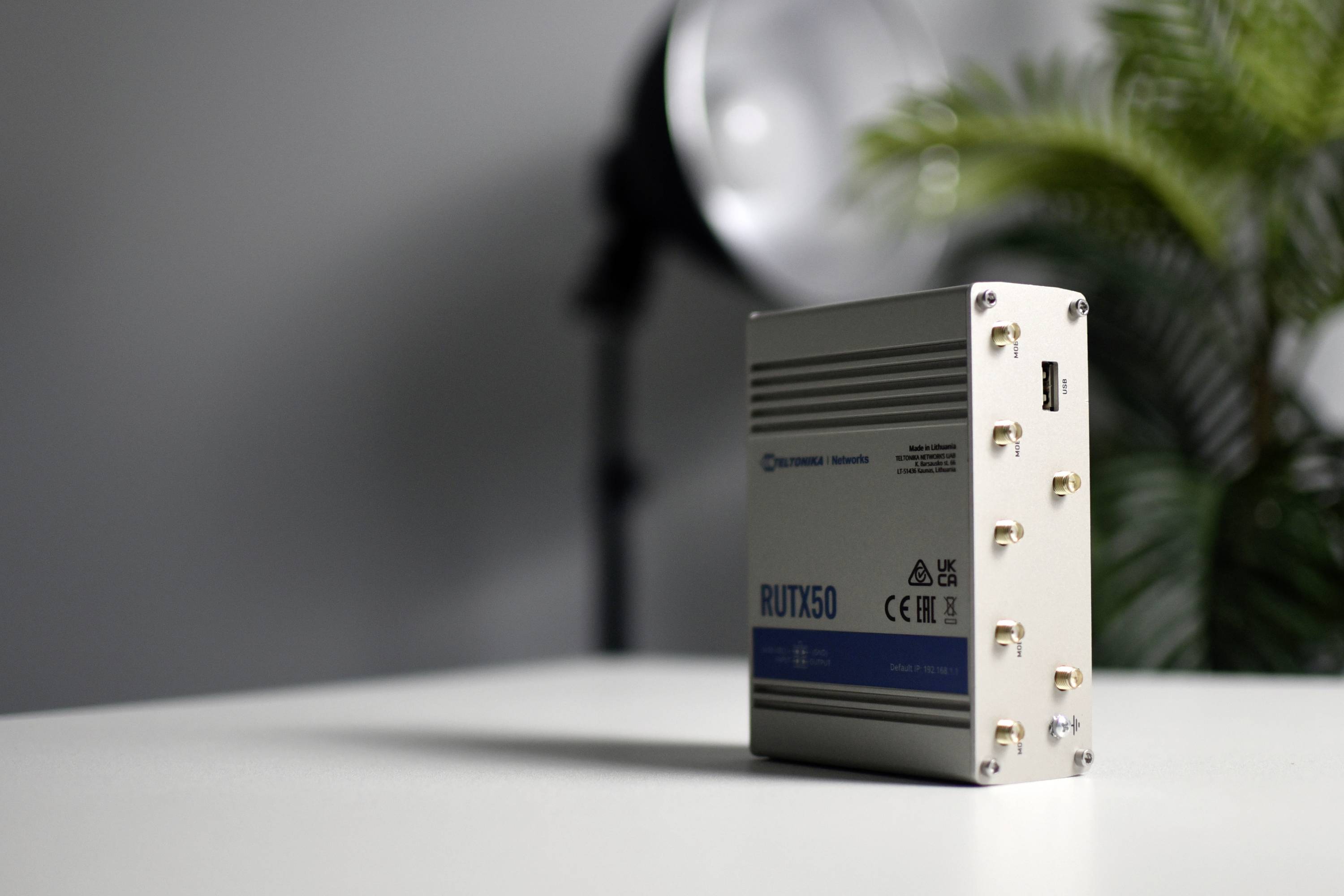 Kleines, silbergraues elektronisches Gerät mit der Aufschrift 'RUTX50' auf einem Tisch, im Hintergrund eine Pflanze und eine Lampe.