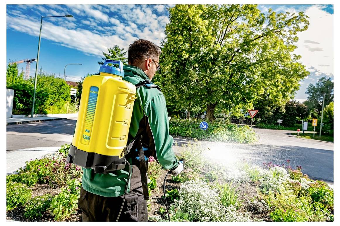 Eine Person nutzt eine Rückenspritze, um einen Garten mit Blumen und Pflanzen in einer hellen, sonnigen Parklandschaft zu bewässern und zu pflegen.