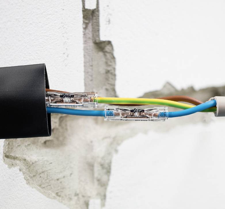 Nahaufnahme einer Wandinstallation: Zwei Kabel mit Klemmen ragen aus einem schwarzen Rohr. Die Wand ist weiß und teilweise offen.