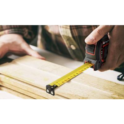 Eine Person misst ein Holzbrett mit einem Maßband. Die Skala zeigt etwa 26 Zentimeter an.