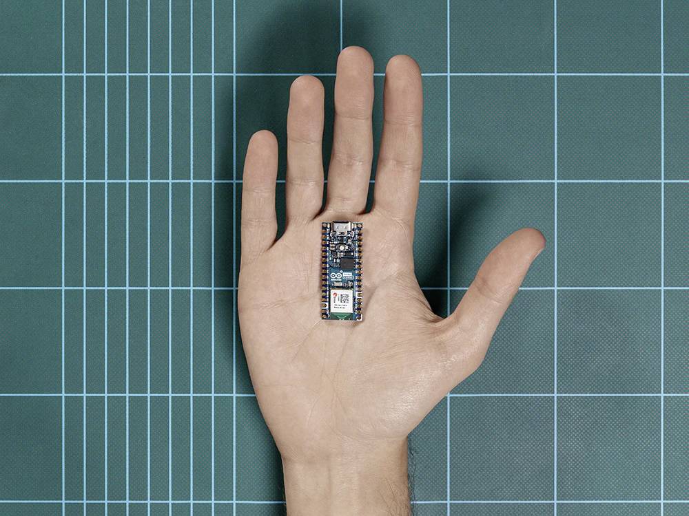 Eine geöffnete Hand zeigt einen kleinen Microcontroller auf einer grünen Gittermatte. Der Microcontroller ist winzig im Vergleich zur Hand.