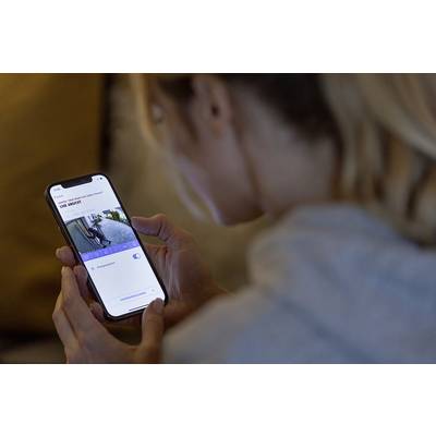 Eine Person betrachtet ein Smartphone, das eine Live-Überwachungskamera zeigt. Die Kamera zeigt den Eingang eines Hauses.