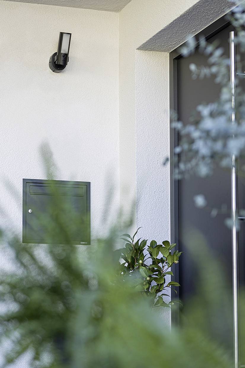 Eine moderne Hauswand mit schwarzem Briefkasten und Außenleuchte, umgeben von Pflanzen. Ein Teil einer Eingangstür ist sichtbar.