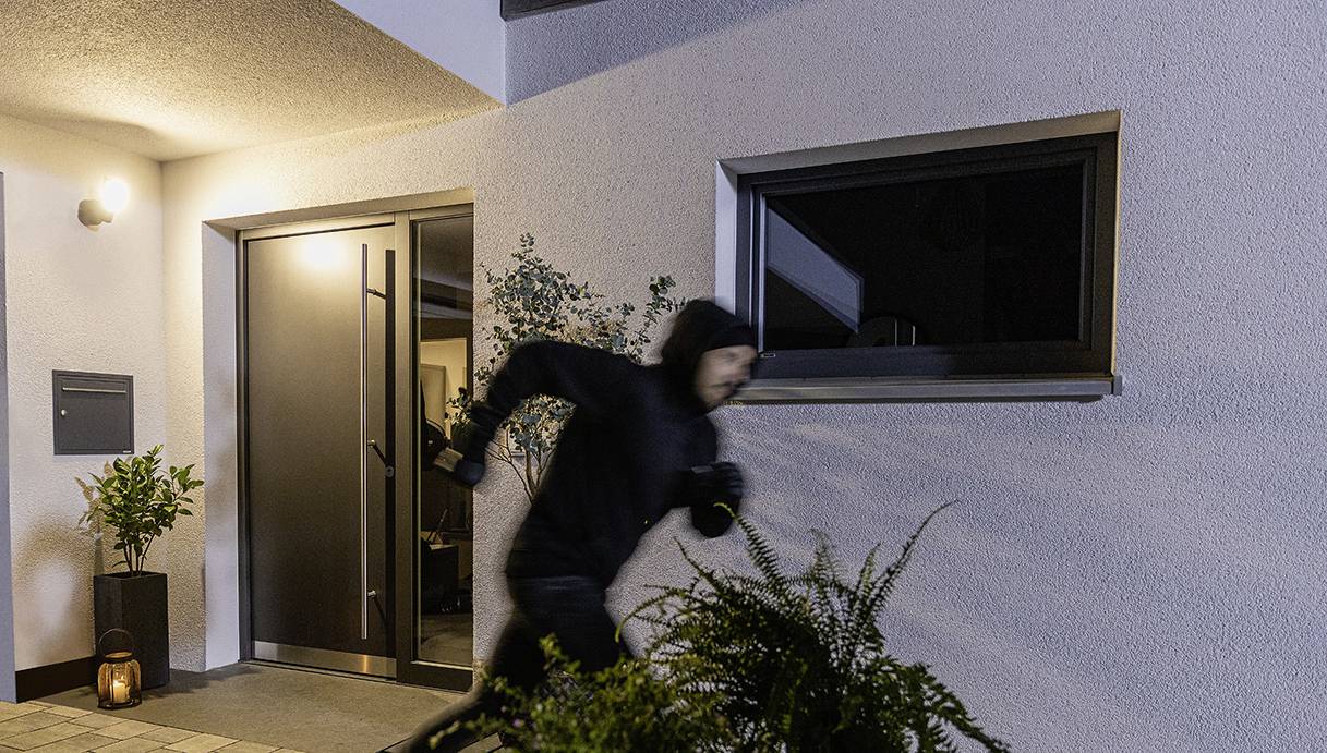 Ein dunkel gekleideter Mensch rennt nachts vor einem Haus mit einer offenen Tür. Die Szene vermittelt Eile oder Flucht.