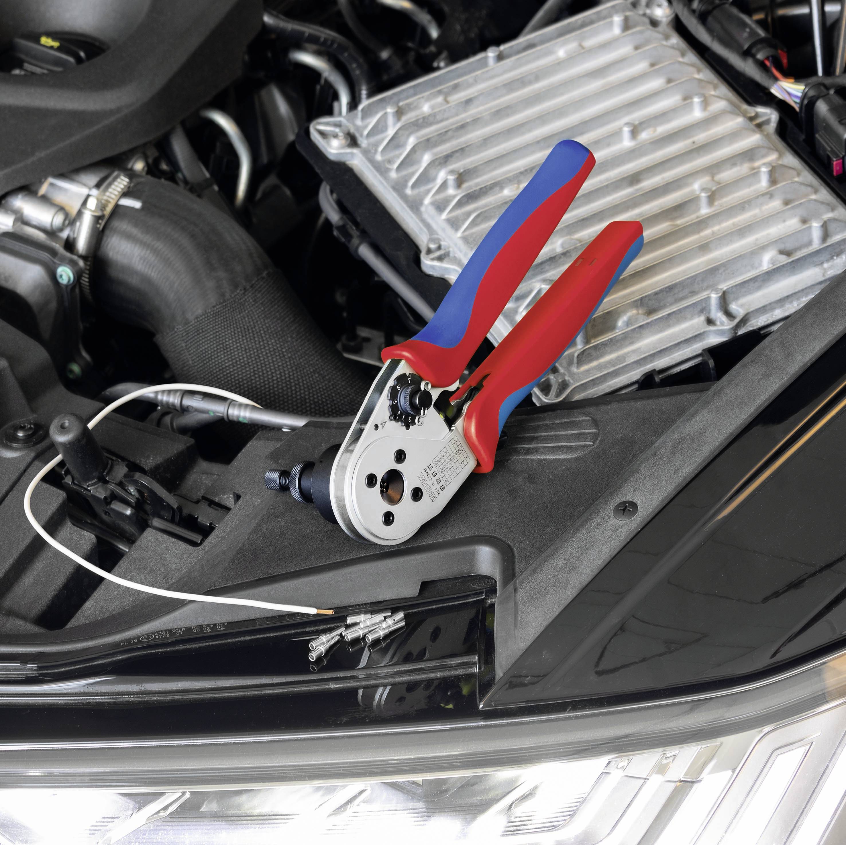 Eine rote und blaue Crimpzange liegt auf einem Autoteil unter einer geöffneten Motorhaube, neben einem abisolierten Kabel.