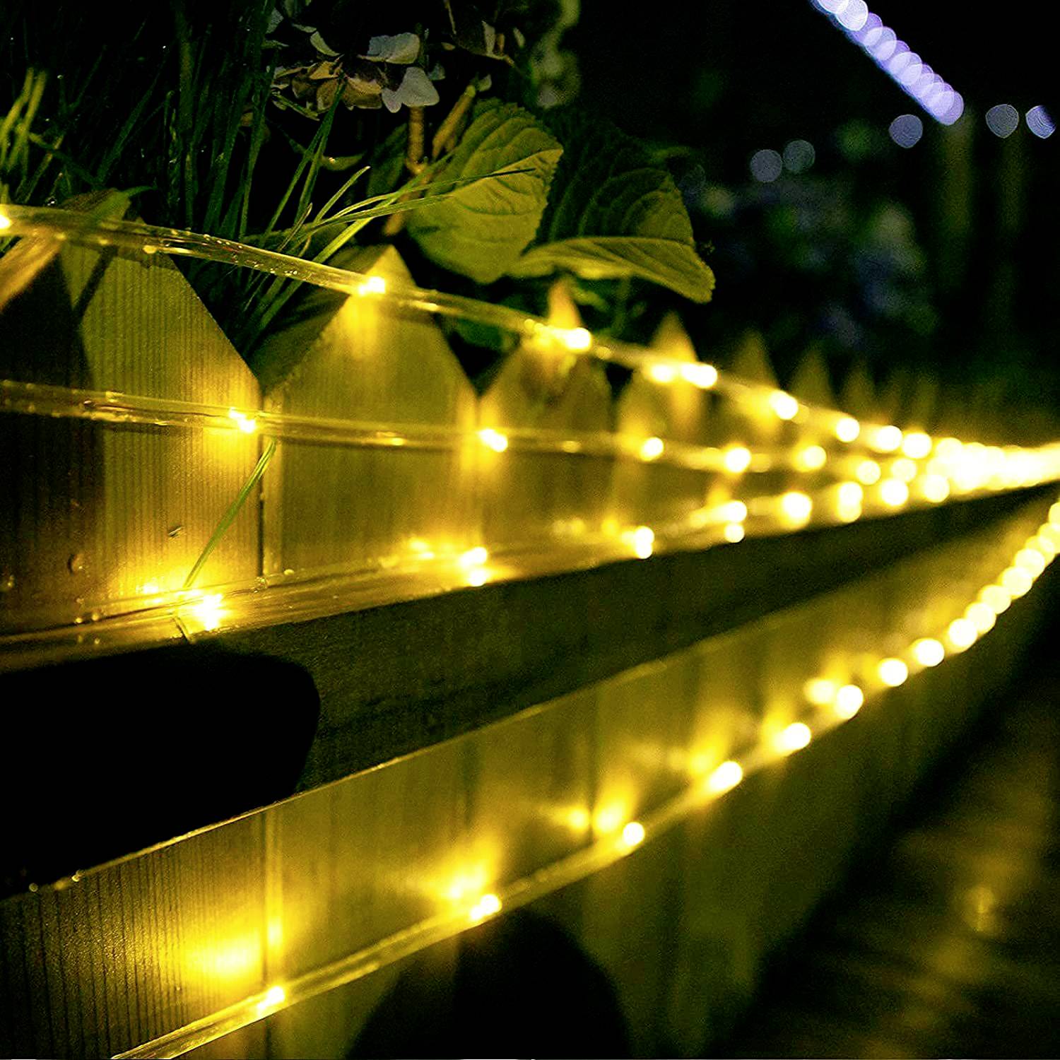Gelbe Lichterketten leuchten auf einem hölzernen Geländer in der Nacht, umgeben von Pflanzen, schaffen eine gemütliche Atmosphäre.