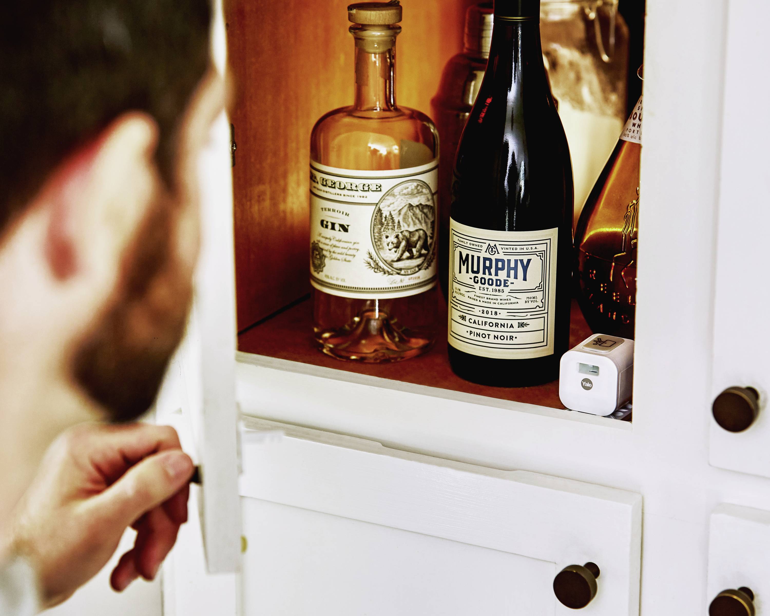 Ein Mann öffnet eine Küchenschranktür und schaut auf mehrere Getränkeflaschen, darunter Gin und kalifornischer Wein.