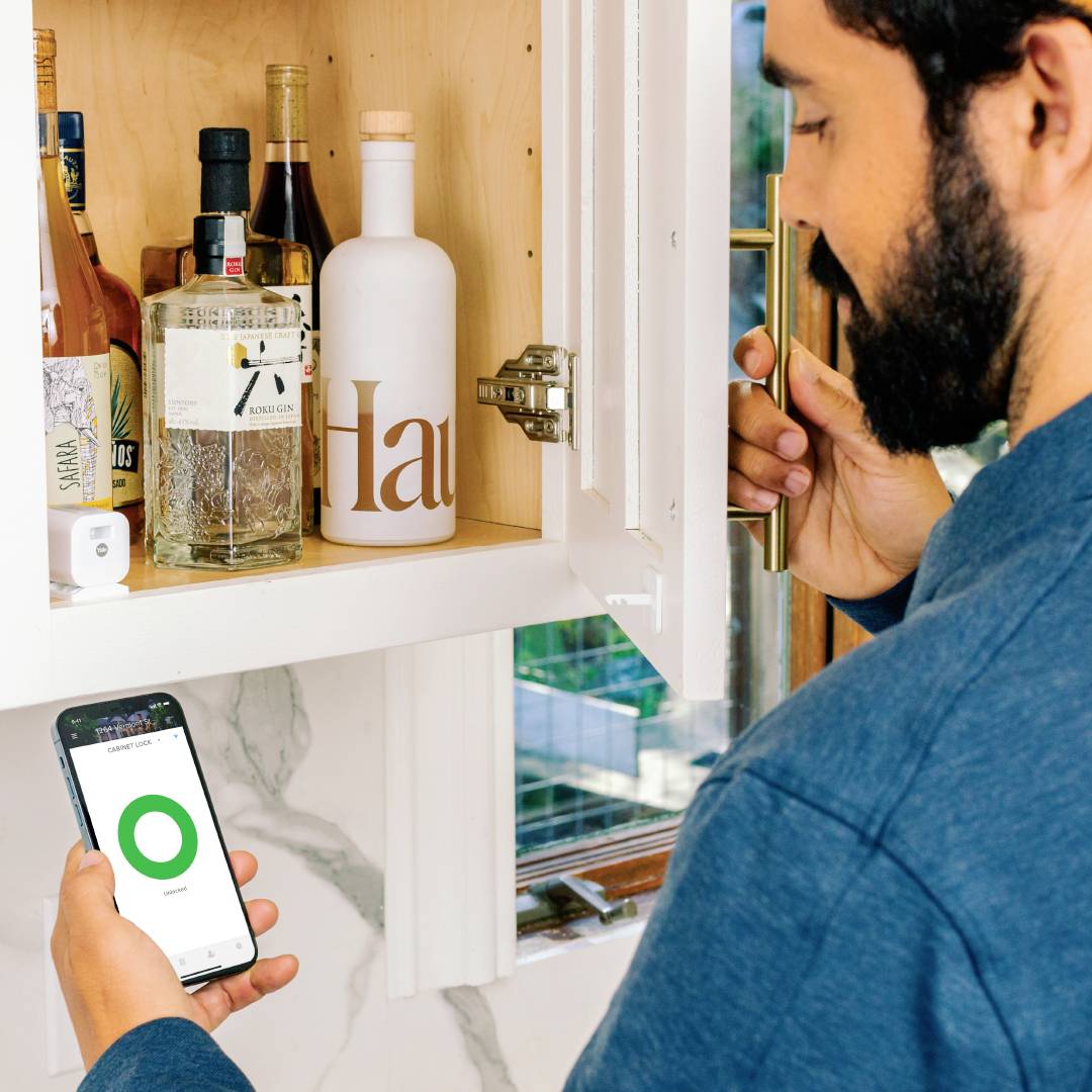 Eine Person scannt mit einem Smartphone einen QR-Code an einer geöffneten Küchenschranktür, hinter der mehrere Flaschen stehen.