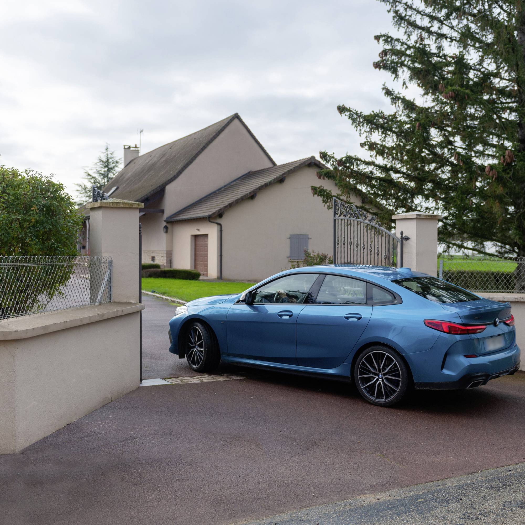 Ein blaues Auto steht auf einer asphaltierten Einfahrt vor einem modernen Haus mit rotem Dach; umgeben von Bäumen und Büschen.