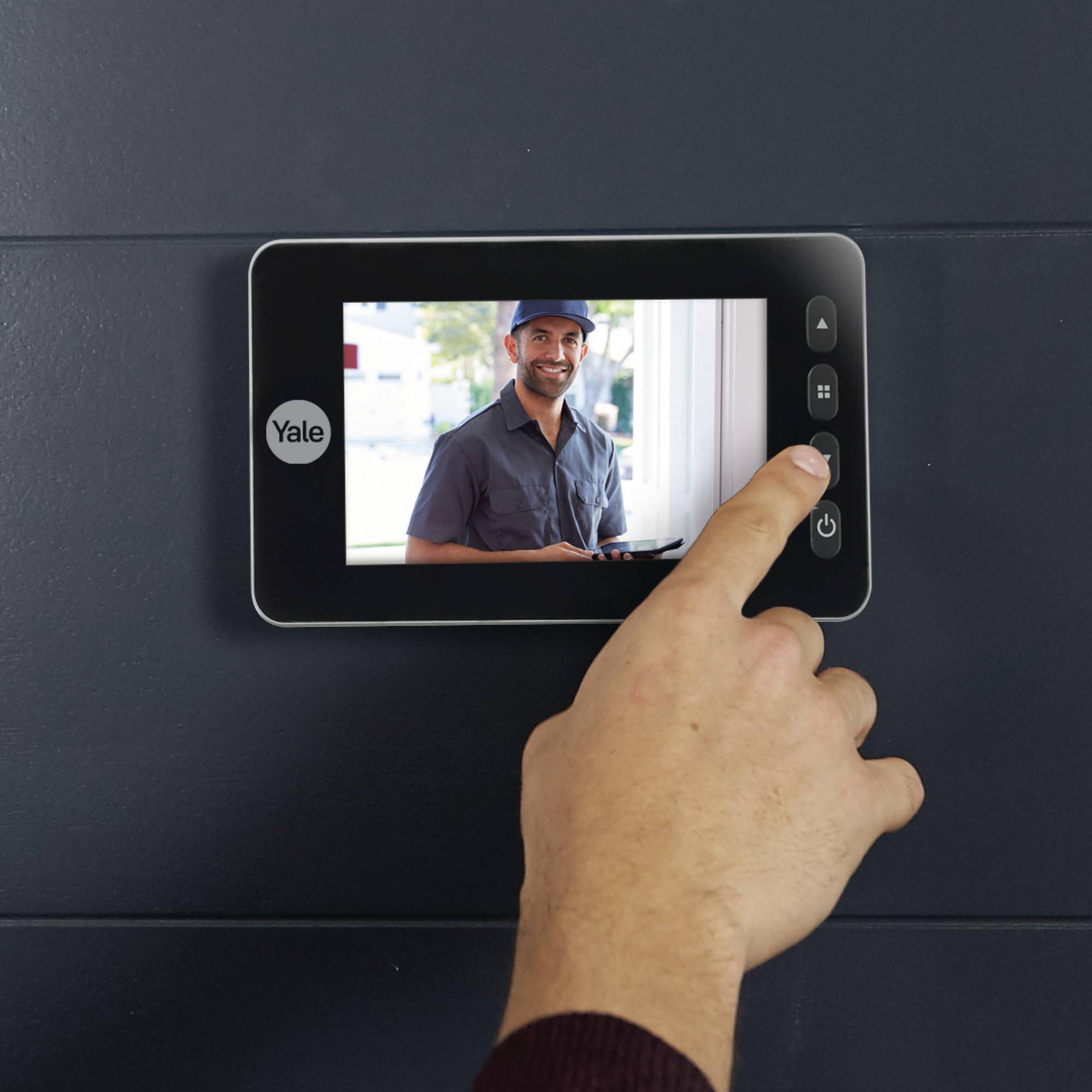 Hand betätigt den Monitor eines digitalen Türspions der Marke Yale mit Videofunktion, der einen lächelnden Zusteller anzeigt.