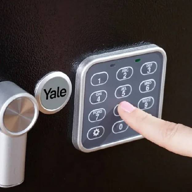 Ein Finger einer Person drückt auf einen Knopf eines digitalen Zahlenschlosses der Marke "Yale" an einer Tür, was einen Sicherheitszugang signalisiert.