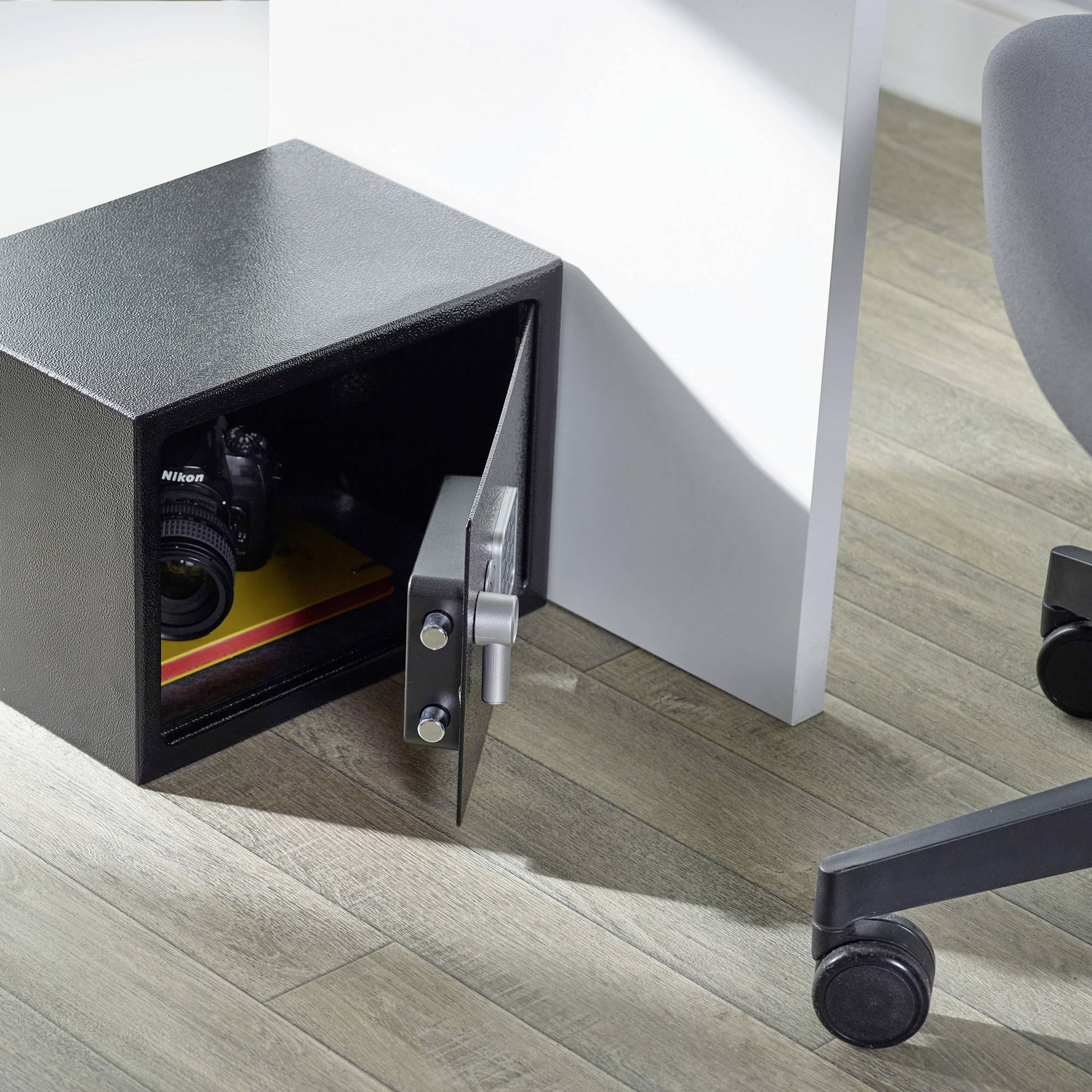 Ein geöffneter schwarzer Safe auf einem Holzboden enthält eine Kamera und ein Buch. Daneben steht ein Bürostuhl.