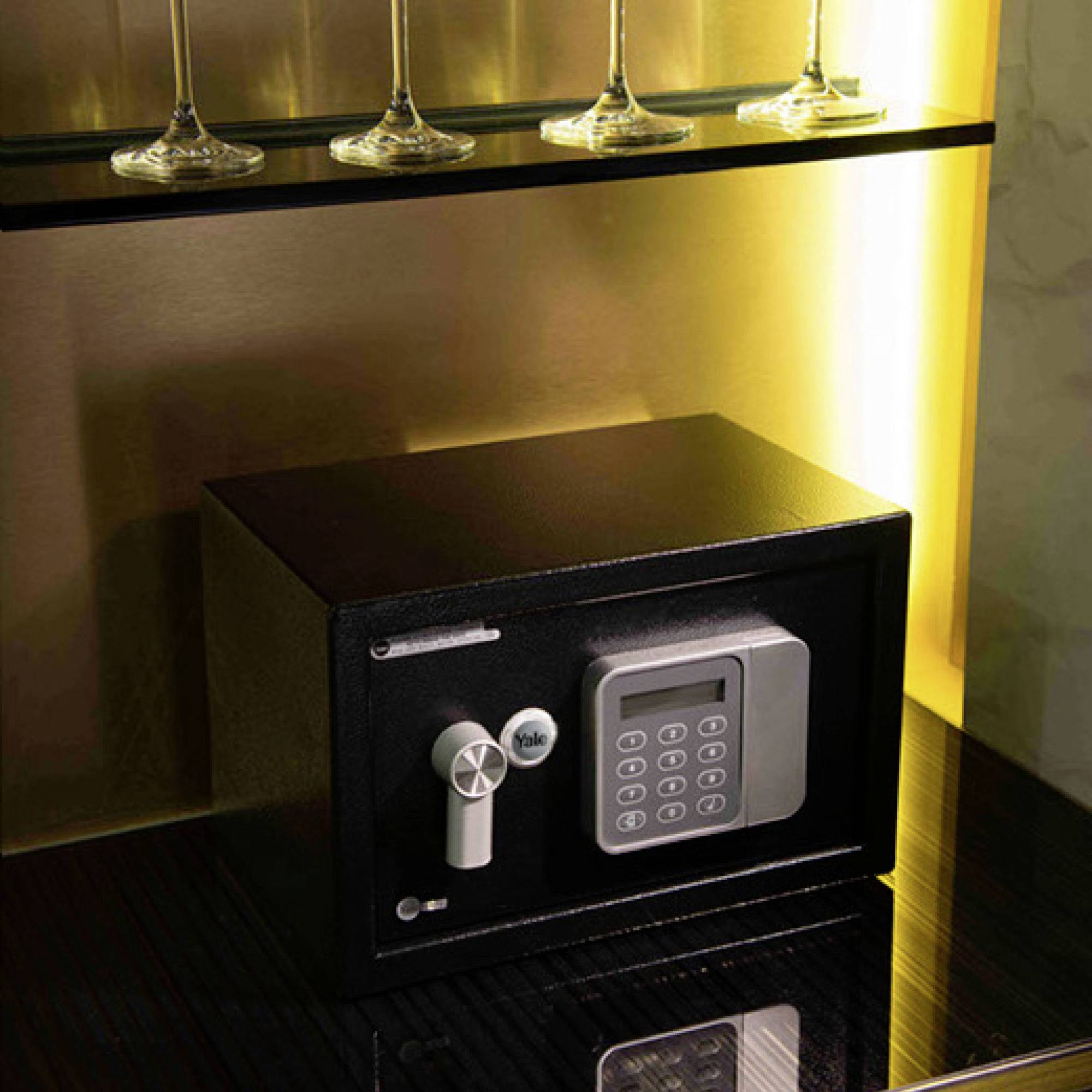 Ein Tresor mit elektronischem Zahlenschloss steht auf einem Holzregal unter einer Reihe von Weingläsern, dezent beleuchtet.