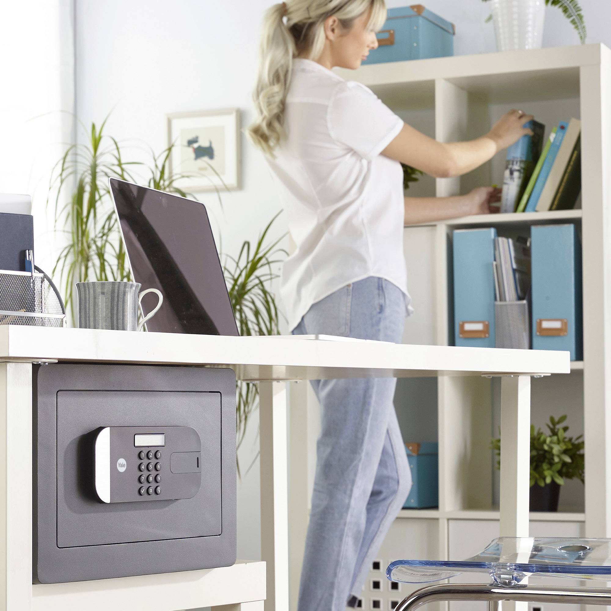 Frau stellt Ordner in ein Regal in einem modernen Büro, Laptop auf dem Tisch und ein Safe montiert an der Tischseite.