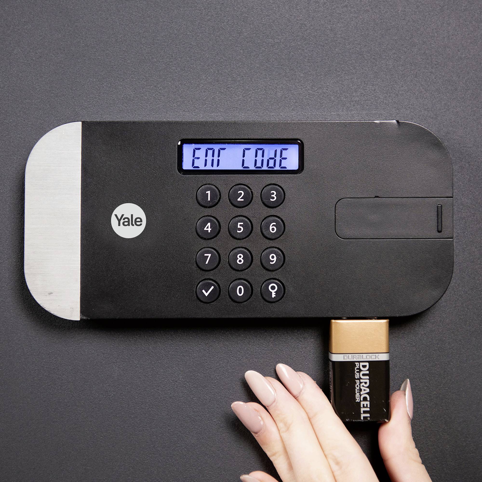 Eine Hand hält eine Batterie, die in die Unterseite eines elektronischen Zahlenschlosses mit der Aufschrift 'Enter Code' eingesetzt ist.