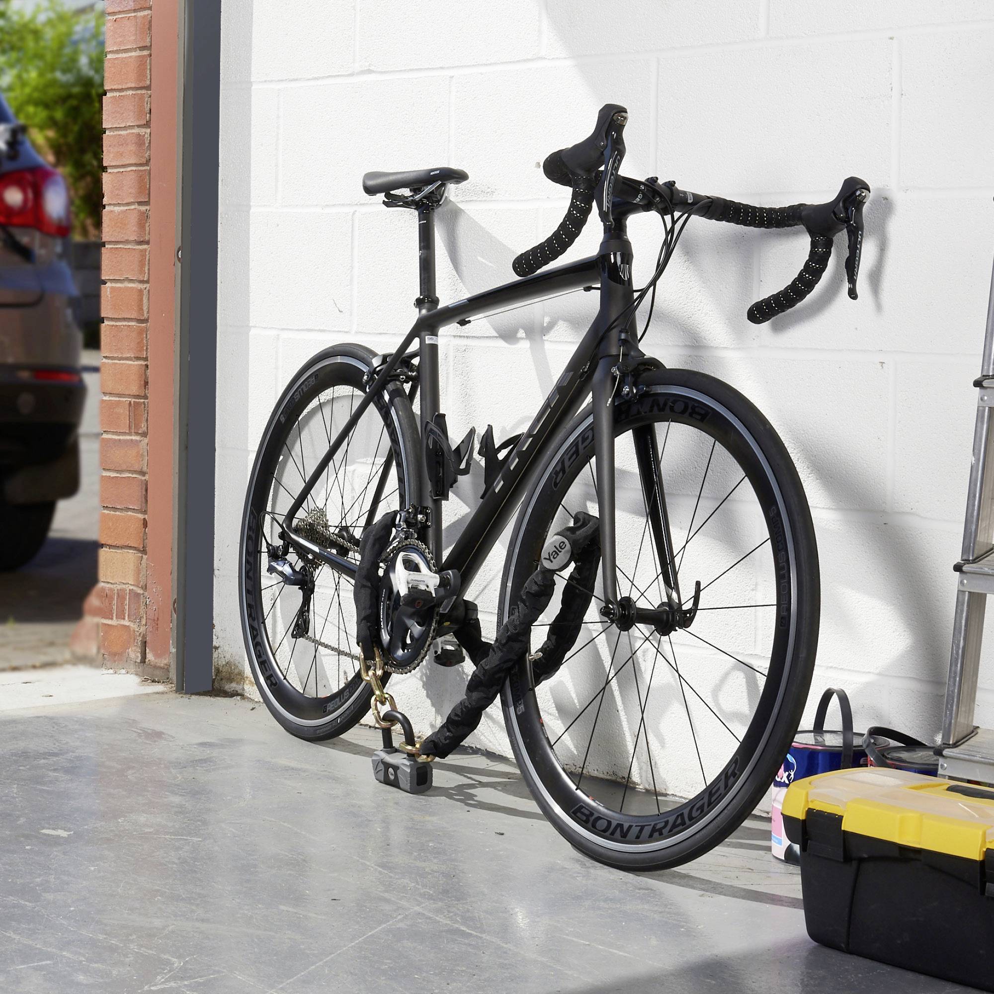 Ein schwarzes Rennrad ist an eine weiße Garagenwand gelehnt und mit einem Schloss gesichert. Rechts steht Werkzeug und ein Karton.