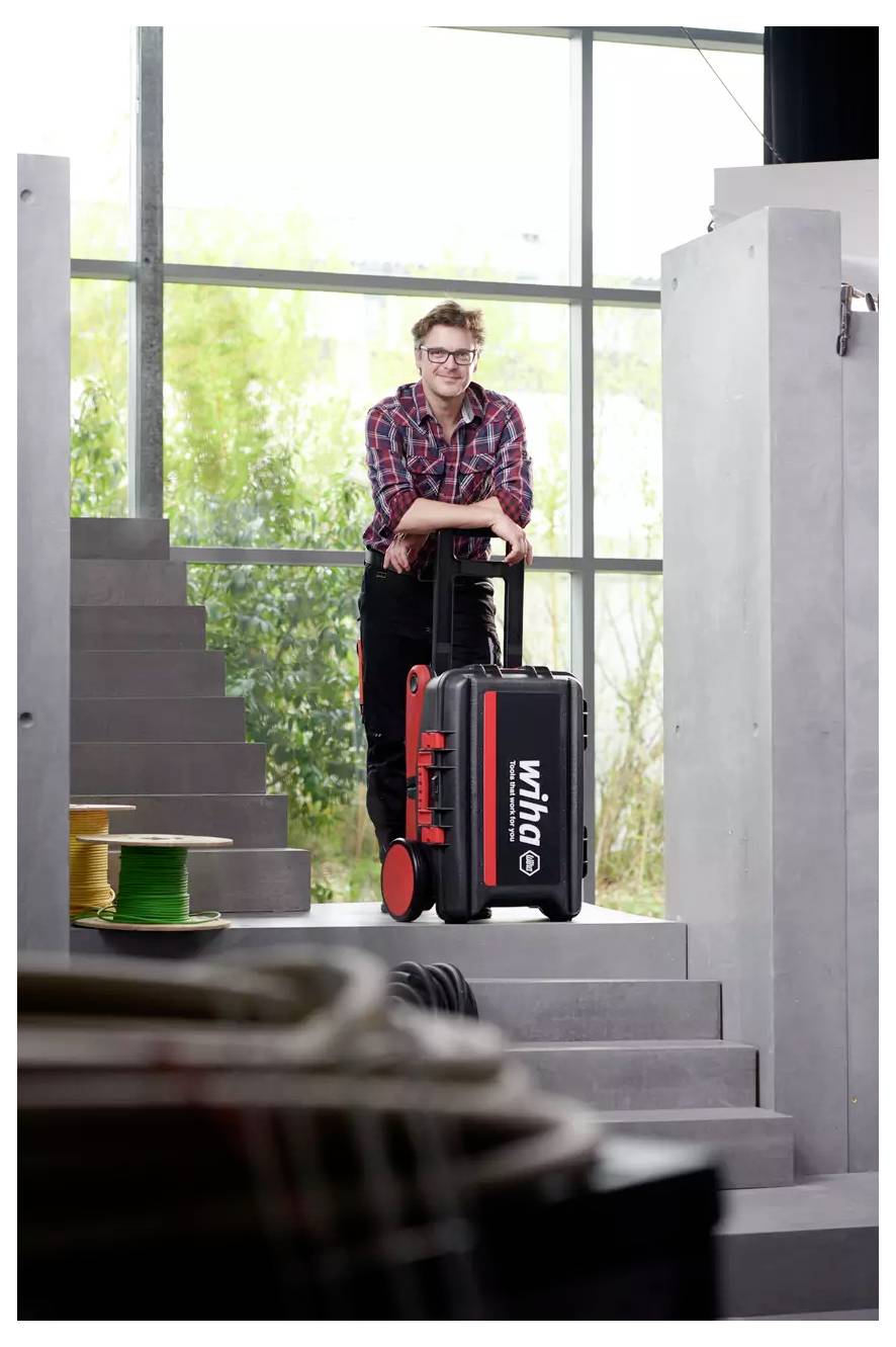 Eine Person in einem karierten Hemd lächelt, während sie sich auf einen Werkzeugkasten mit Rollen lehnt, der mit „wiha" beschriftet ist, und auf einer Treppe in einer hellen, industriellen Umgebung steht.