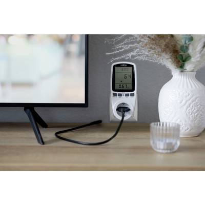 Ein Strommessgerät zeigt Verbrauchsdaten an einer Steckdose neben einem Fernseher. Daneben steht eine dekorative Vase.