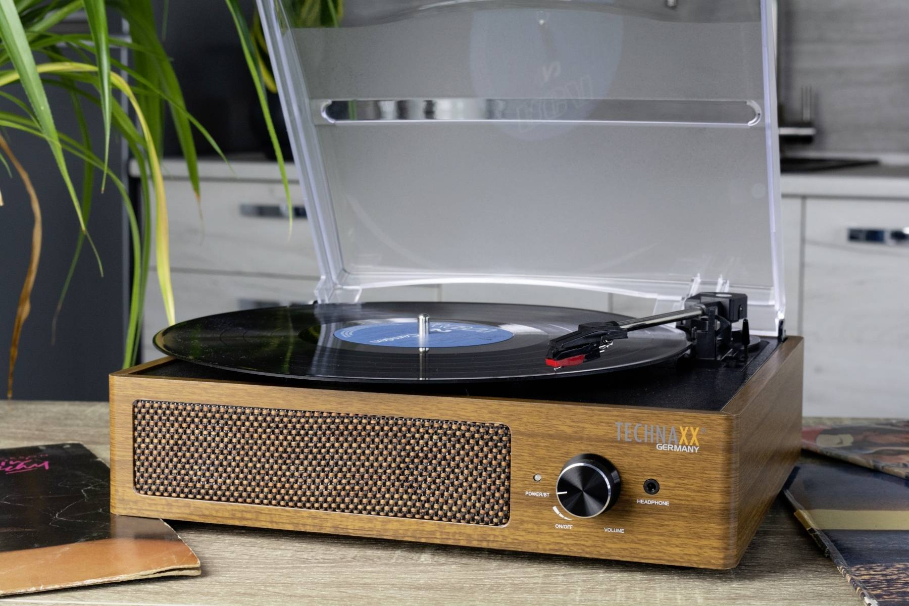 Plattenspieler aus Holz mit aufgelegter Schallplatte, umgeben von weiteren Schallplatten. Im Hintergrund ist eine Pflanze sichtbar.