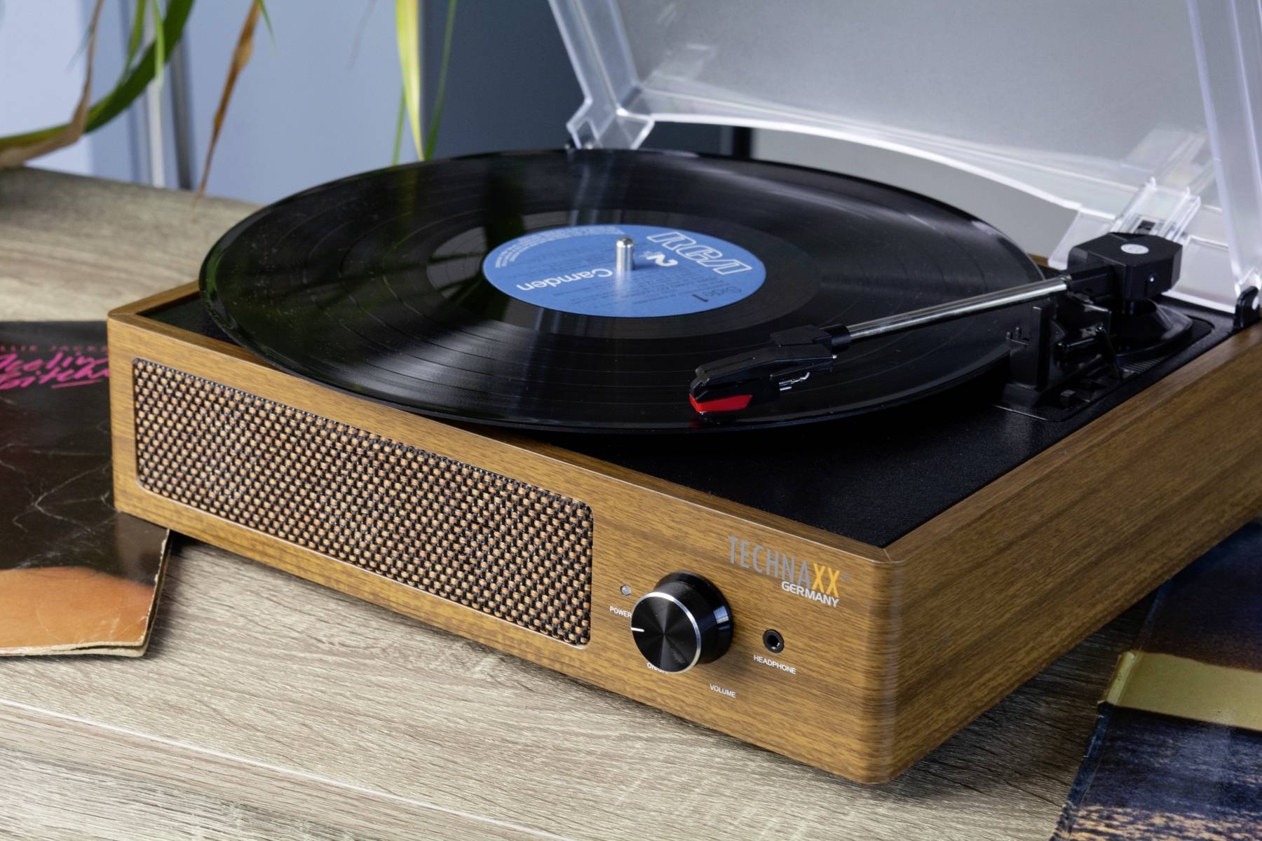Plattenspieler in Holzoptik mit schwarzer Vinyl-Schallplatte auf einem Tisch; nebenbei sind zwei weitere Schallplattenhüllen sichtbar.