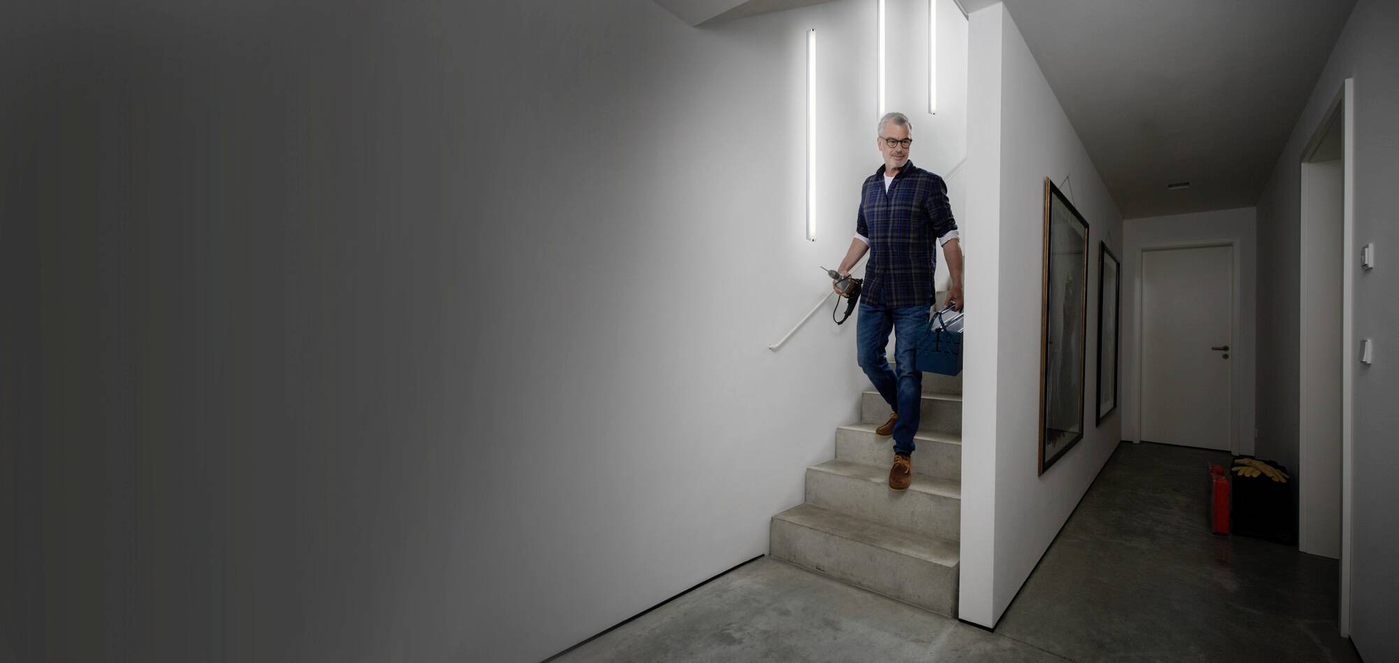 Ein Mann geht eine Treppe in einem modern beleuchteten, weißen Flur hinunter. Er trägt Freizeitkleidung und hält etwas in der Hand.