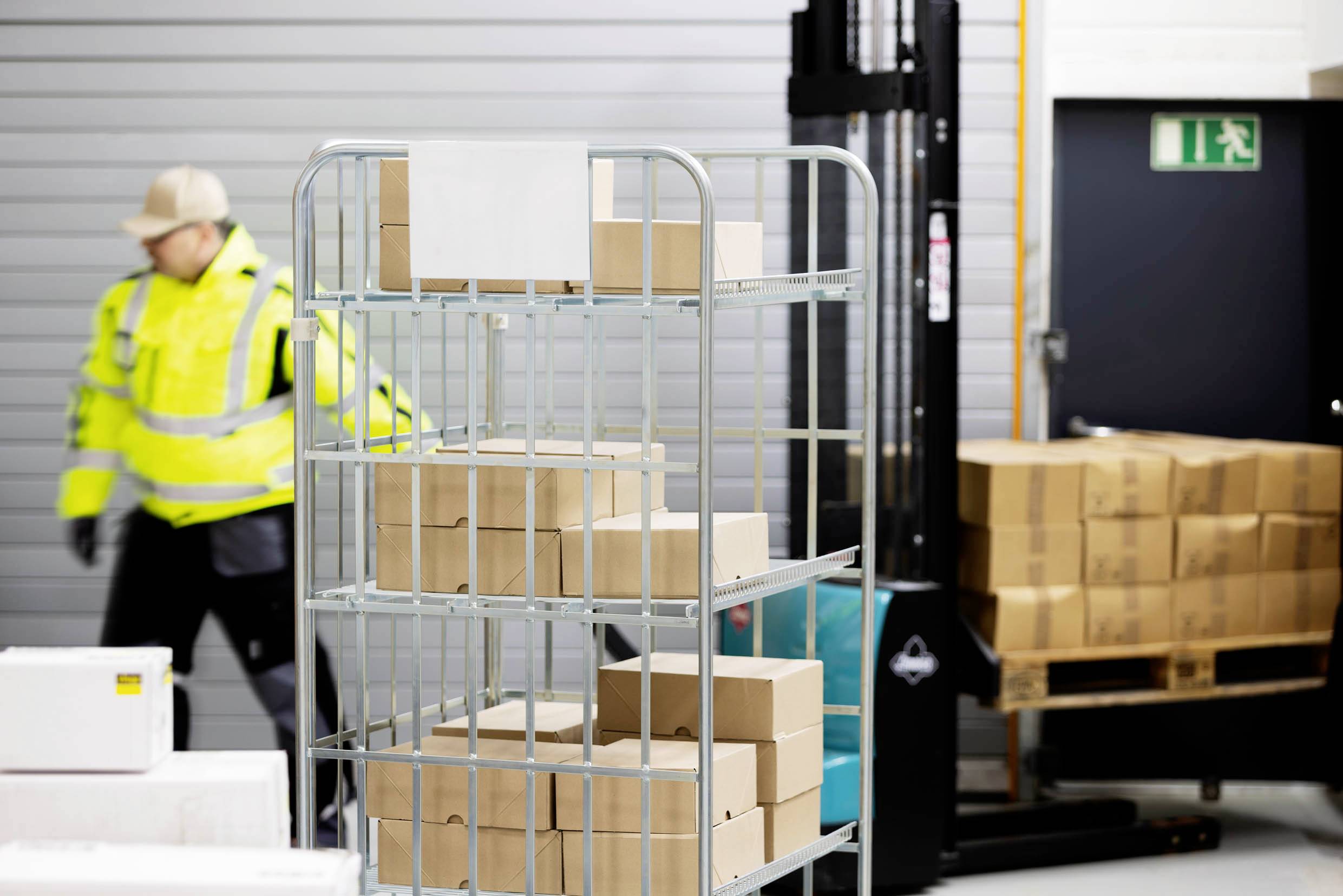 Eine Person in Arbeitskleidung schiebt einen Wagen mit Kisten in einem Lagerhaus; ein Gabelstapler steht daneben.