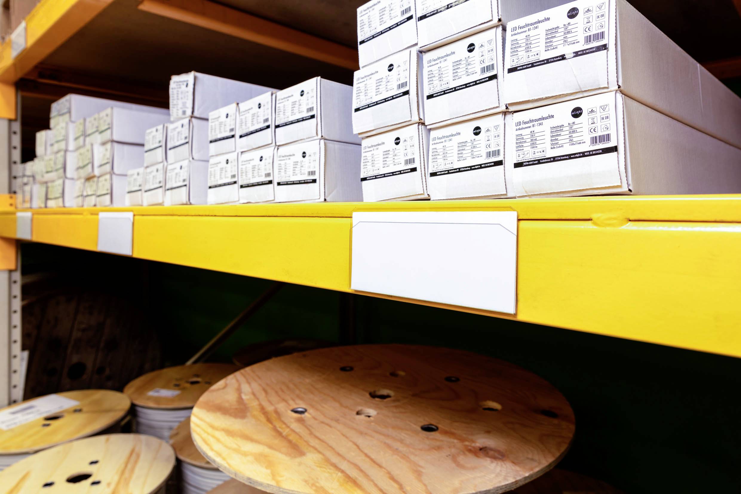 Regal mit weißen Kartons beschriftet mit technischen Details, darüber Holztrommeln mit Kabeln. Lagerumgebung mit Regalen.