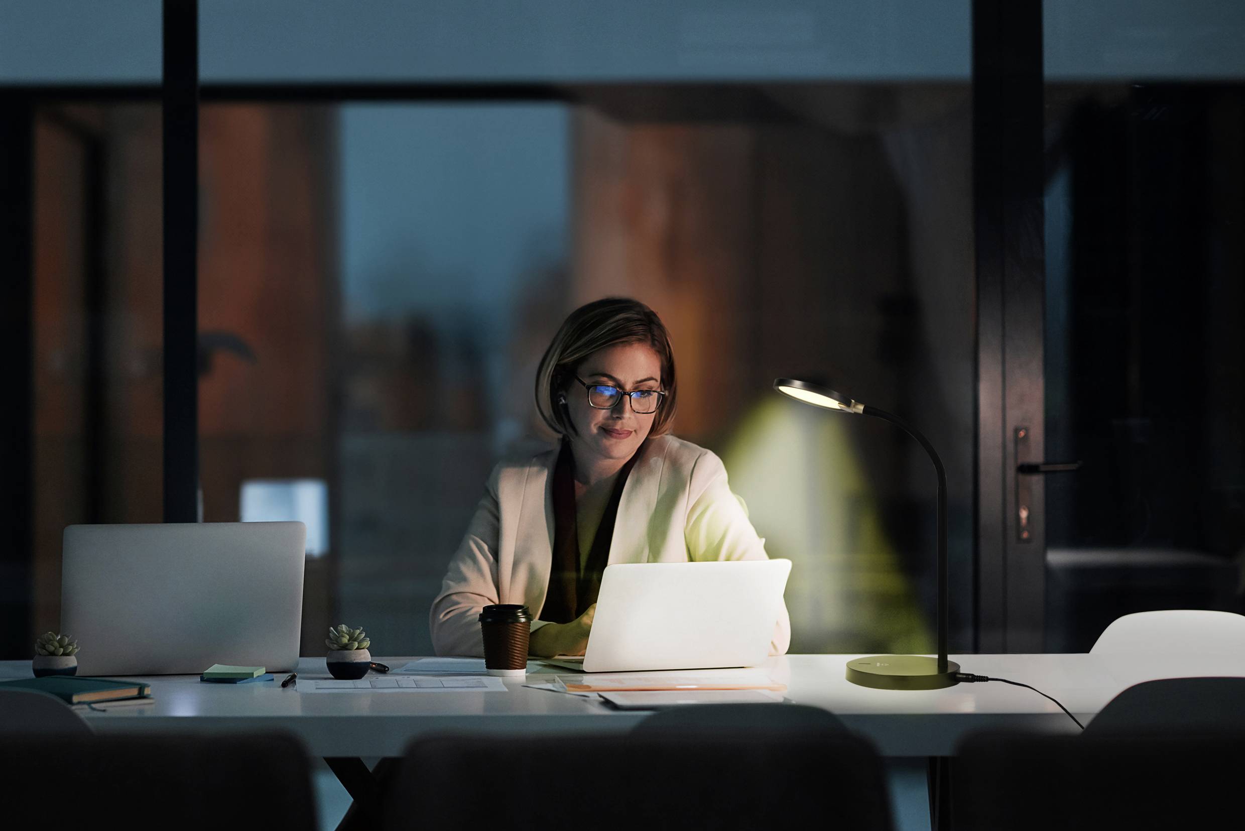 Eine Frau arbeitet abends an einem Laptop in einem Büro. Der Raum ist durch eine Schreibtischlampe beleuchtet.