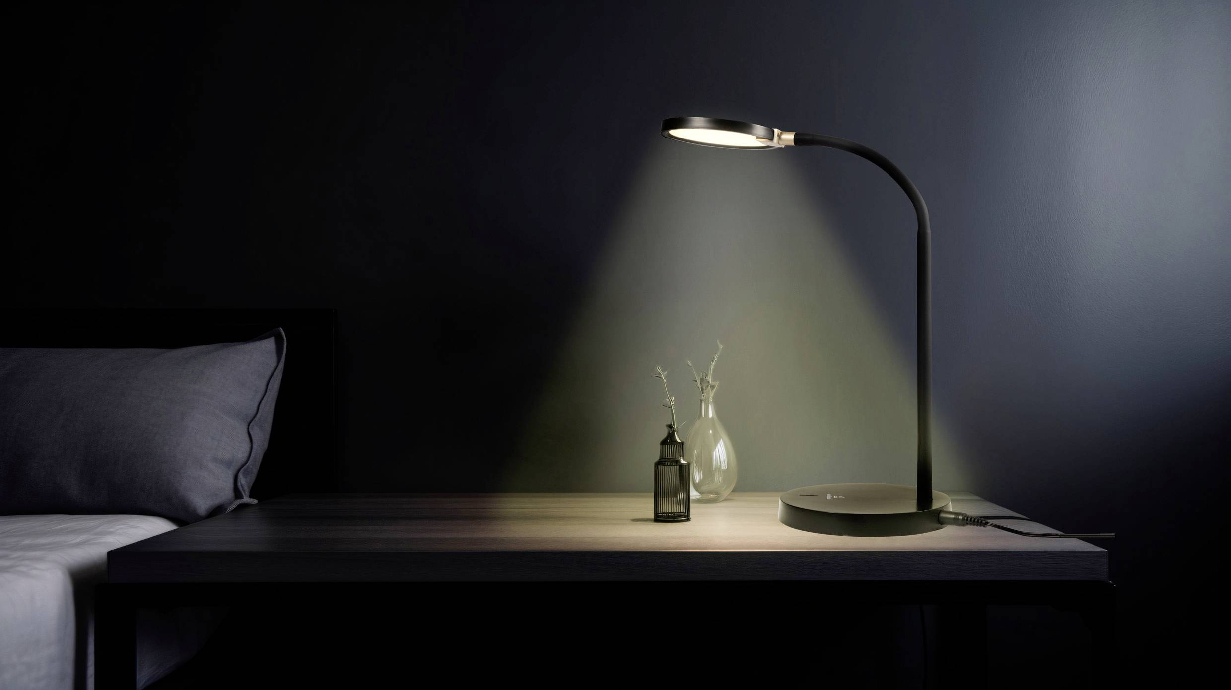 Nachttisch mit eingeschalteter Lampe, die sanftes Licht auf einen Tisch mit kleinem Blumenstrauß und Flasche wirft.