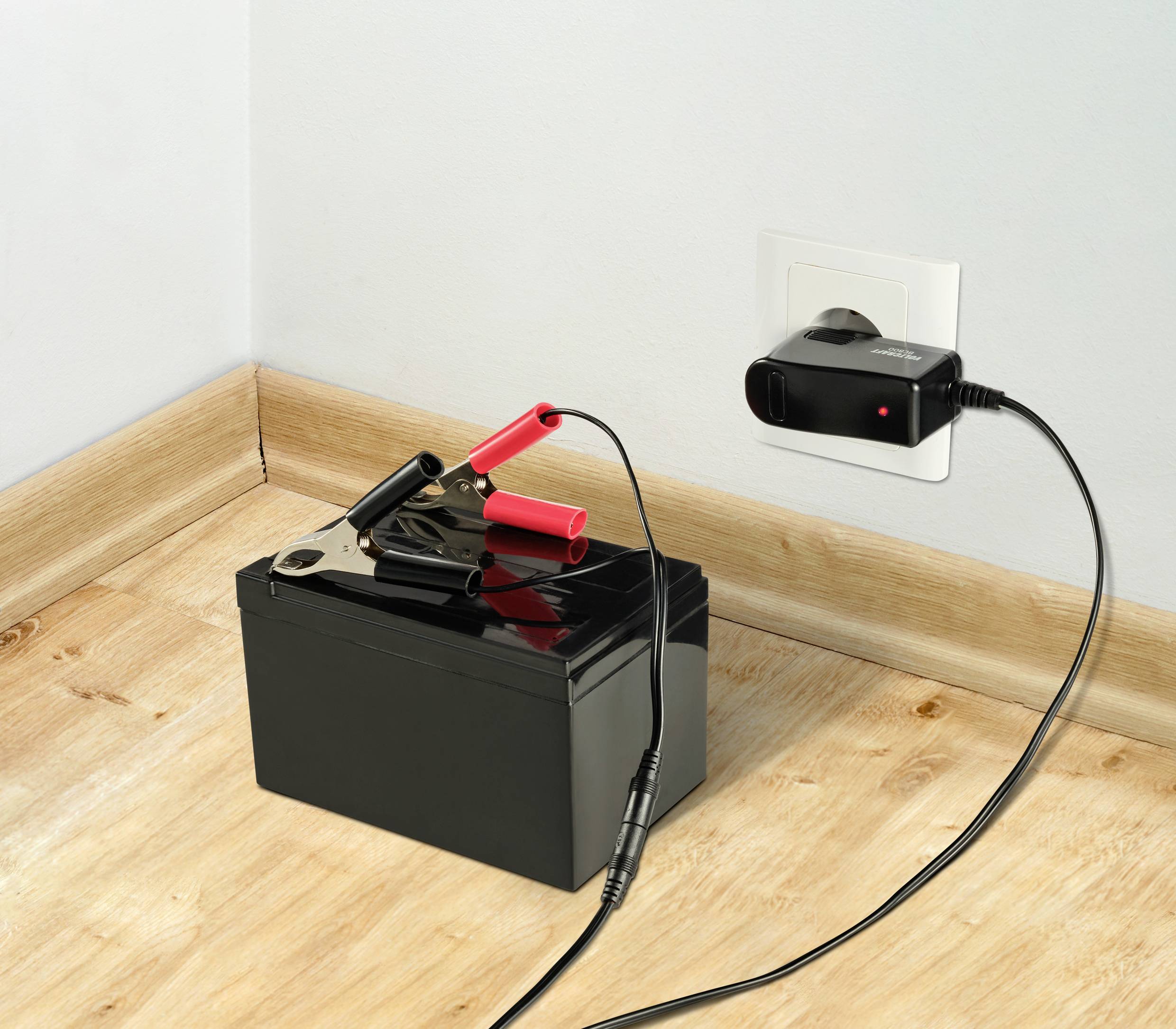 'Eine Autobatterie mit angeschlossenen Klemmen wird über ein Ladegerät, das in eine Steckdose eingesteckt ist, geladen.'